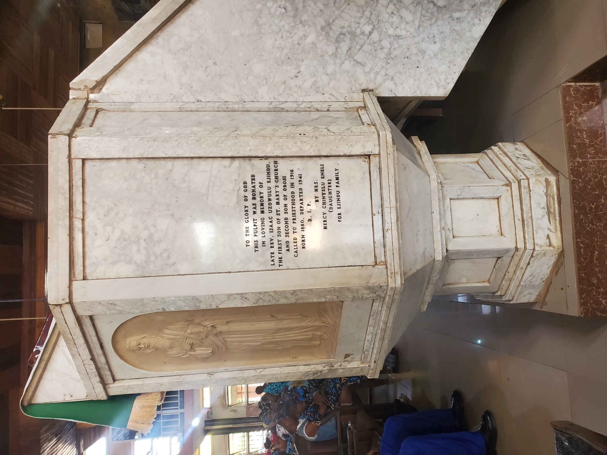 The image shows a white marble pulpit in Nkpor, Nigeria. The pulpit is inscribed with the following text: "TO THE GLORY OF GOD, THIS PULPIT WAS DONATED IN LOVING MEMORY OF LATE REV. ISAAC UZOWULU EJINDU. THE FIRST SON OF ST. MARY'S CHURCH AND SECOND SON OF OBOSI CALLED TO PRIESTHOOD IN 1916 BORN 1880, DEPARTED 1941 R. I. P. BY MRS. MERCY CHINYELU ENELI (DAUGHTER) FOR EJINDU FAMILY". A carved image of a man holding a book is visible. People are seated in the background.