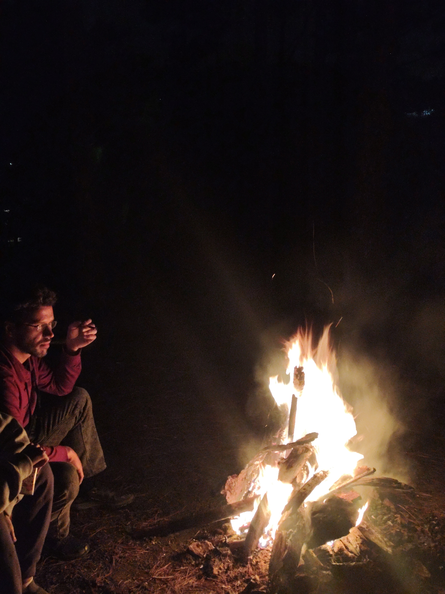 The image portrays a nighttime outdoor scene dominated by a brightly burning bonfire. A man with short, dark, curly hair, glasses, and a beard is seated on the left side of the frame, illuminated by the fire's glow. He is wearing a red long-sleeved shirt and dark pants, with his right hand raised to his mouth, holding what appears to be a pipe or cigar. Partially visible beside him, in the bottom-left corner, are the legs and lower torso of another person, clad in dark pants and a light-colored hooded top.
The central focus is the large bonfire, actively burning with vivid orange and yellow flames that cast smoke upwards. Logs and branches are visible within the fire. A stick, possibly holding a food item like a marshmallow, extends into the flames, suggesting roasting. The ground surrounding the fire appears to be a mix of dirt and dry foliage.
The scene depicts a group of individuals gathered around a campfire at night, likely for a social event or camping. The visible man's posture and expression suggest a moment of relaxation and contemplation as he observes the fire. The atmosphere is intimate and cozy, with the bonfire providing the sole significant light source against the dark background. Faint distant lights are barely discernible in the darkness, hinting at a remote or wilderness setting. No text is visible in the image.