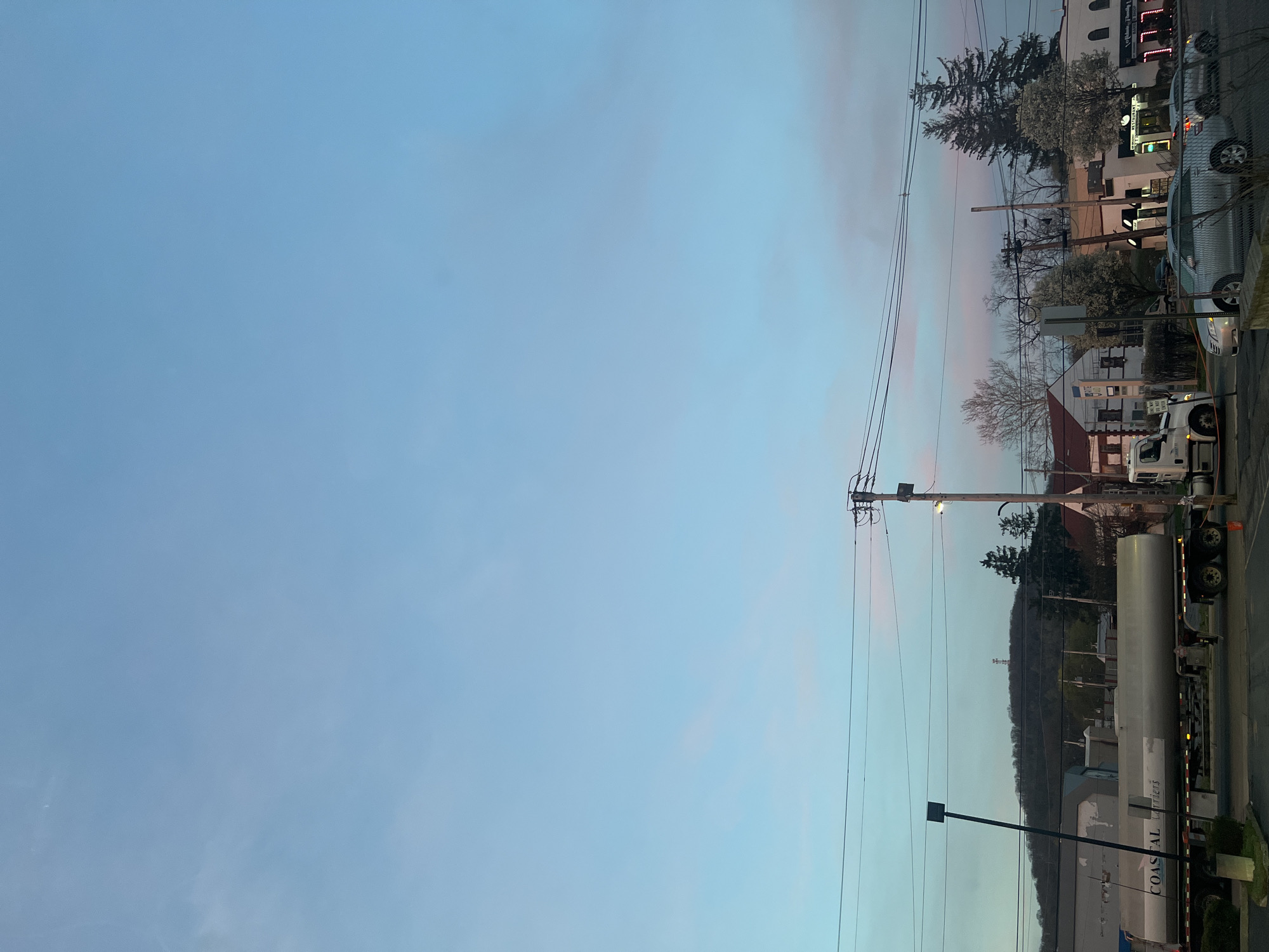 The image captures an urban scene in the City of Newburgh, United States of America, at dusk. Predominantly, the sky is a gradient of blue with subtle pink hues. On the right side of the frame, a street scene is visible, including utility poles with power lines, buildings, and a variety of parked vehicles. A large tanker truck with the word "COASTAL" on it is a prominent object. There is also a hill behind the building. A street light post is situated near the tanker truck.