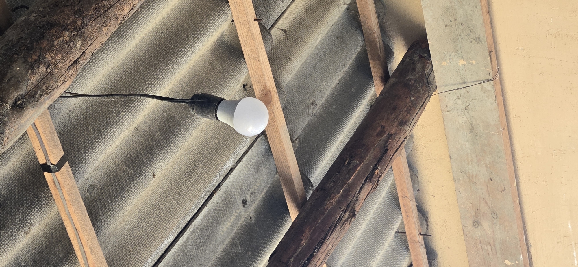 A white LED light bulb hangs from a black cord, suspended from a wooden beam supporting a corrugated roof. The roof appears to be made of asbestos cement sheeting. Wooden beams, some weathered and dark, are visible supporting the roof structure. The overall scene suggests an outdoor or un finished interior space, possibly a shed or agricultural building. No people or specific identifiable locations within Tel, Azerbaijan are visible or discernible from the image. The lighting is natural, casting shadows and highlighting the textures of the materials.