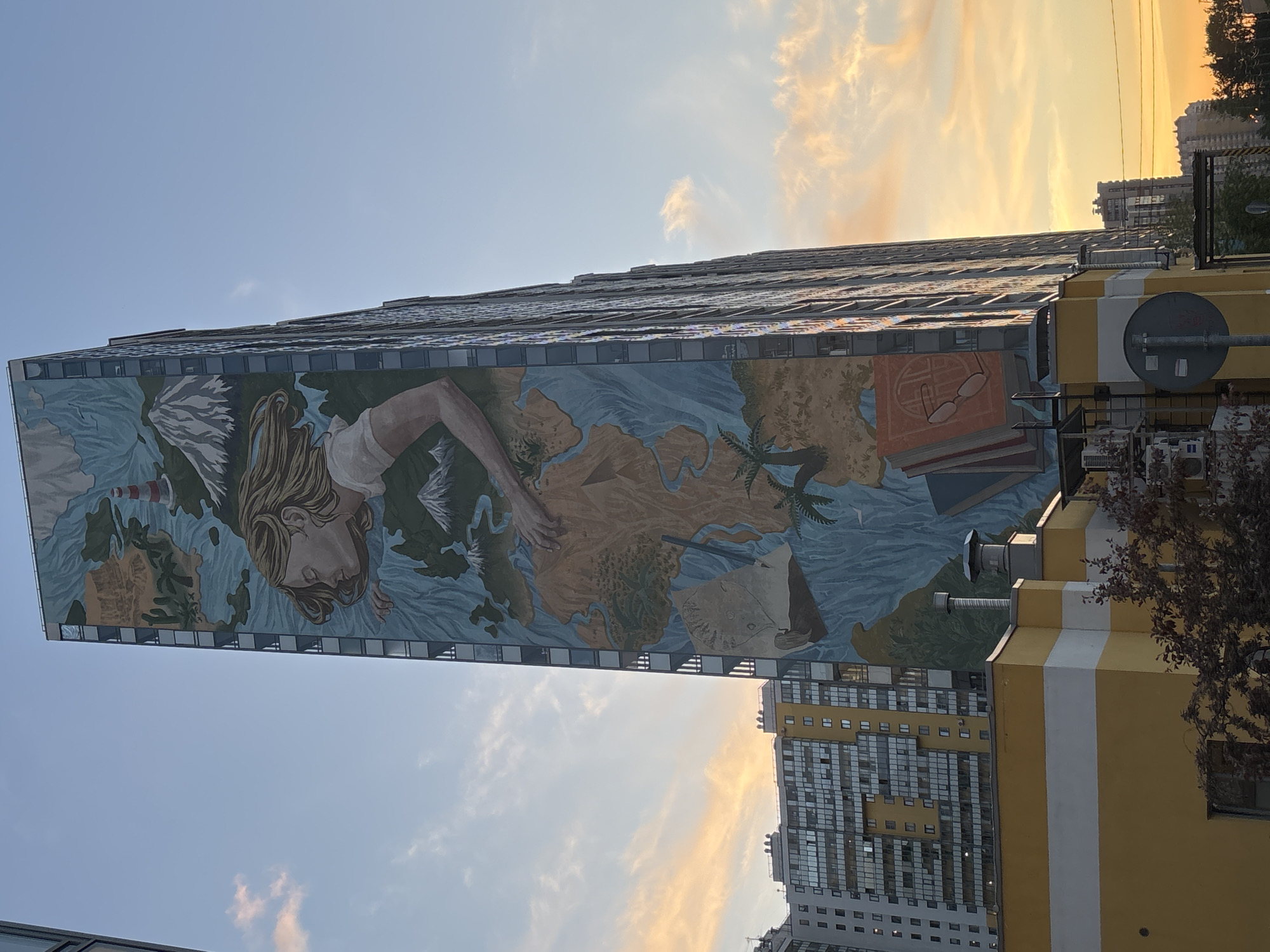 The image displays a striking, multi-story mural covering the side of a tall urban building, captured during the golden hour as the sun sets. The sky on the right side of the frame is bathed in warm orange and yellow hues, fading into a softer blue with scattered white clouds towards the left.
The mural itself is the central subject, depicting a large female figure with long, flowing blonde hair. Her form appears intertwined with a vast, blue body of water, which is interspersed with brown and green landmasses, resembling a world map. Within this intricate landscape, various elements are visible: a white lighthouse on the far left, several palm trees on the depicted land, and a classic sailing ship with a white sail near the bottom center. Towards the upper right of the mural, a stack of books is shown, topped by a pair of eyeglasses, suggesting themes of knowledge or exploration.
The building adorned with the mural is tall and modern, with windows visible along its upper edge above the artwork. In the background, another high-rise building with numerous windows can be partially seen. To the right, a smaller, yellow building with prominent white vertical stripes stands, featuring some utility fixtures and partial views of tree branches.
The overall scene combines art and architecture within an urban setting, conveying a sense of global connection, discovery, and wisdom through its symbolic imagery. No discernible text is visible anywhere in the image.