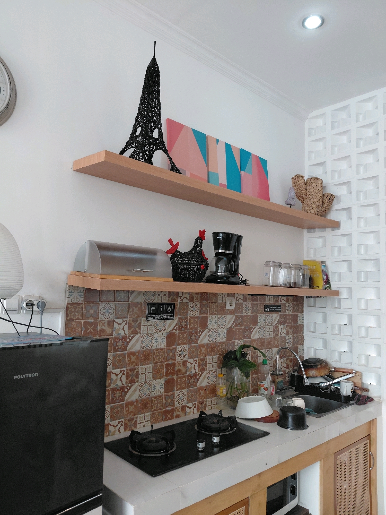 This image depicts a section of a compact kitchen. On a white wall, two light-colored wooden shelves are mounted. The top shelf holds a black Eiffel Tower replica and a piece of abstract art with pink, peach, and teal geometric shapes. To the right, on the same shelf, are two woven basket-like decorative items, one resembling a cactus. The lower shelf displays a clear bread box, a black chicken figurine, a black coffee maker, three glass jars with lids, and a yellow book.
Below the shelves, a backsplash of patterned tiles in brown, beige, and grey hues is visible. A black gas stovetop with two burners is on the white countertop to the left. To the right of the stovetop is a kitchen sink with a faucet. Various items are present around the sink, including a glass vase with a plant, bottles of liquid, a white bowl, a black pot, a white mug, and some kitchen utensils. A black refrigerator with the "POLYTRON" logo is situated to the left of the stovetop. The kitchen cabinets underneath the countertop are made of light wood with woven rattan inserts on the doors. In the background, a white ventilation brick wall is visible. The lighting suggests an indoor environment, likely during the daytime. The location is identified as Batu, Indonesia.
