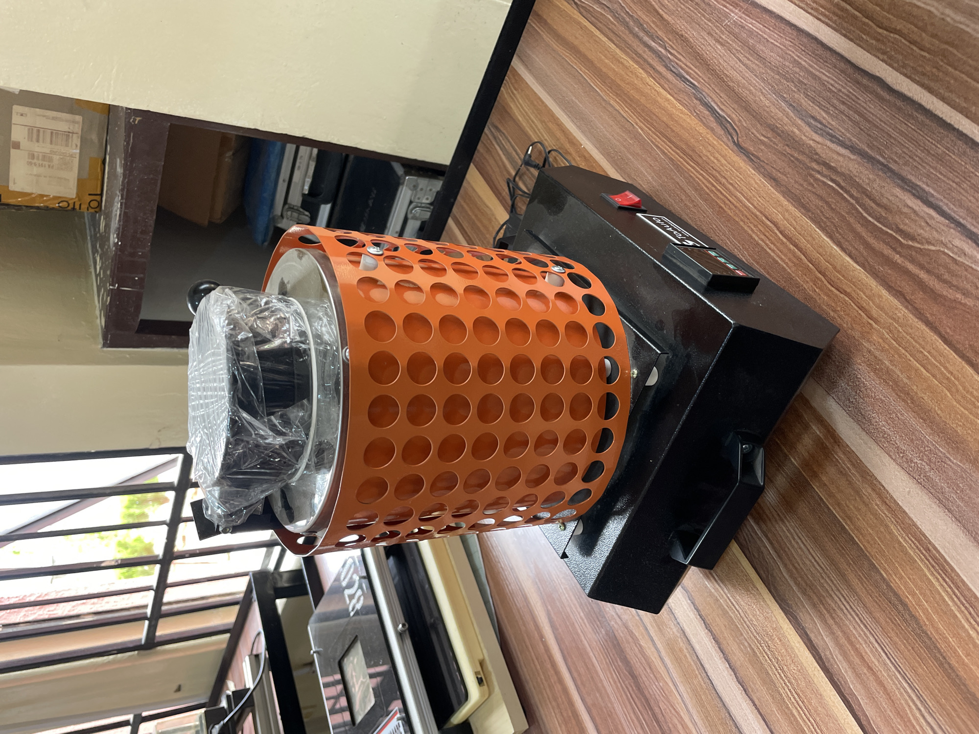 The image shows a piece of machinery with an orange, cylindrical, perforated guard surrounding a cylindrical component wrapped in plastic. The machinery is primarily black and has a red power switch. It rests on a surface with a wood grain pattern. In the background, there are glimpses of an outdoor environment and other equipment. The location is not specified, but the city is Afaka, Nigeria.