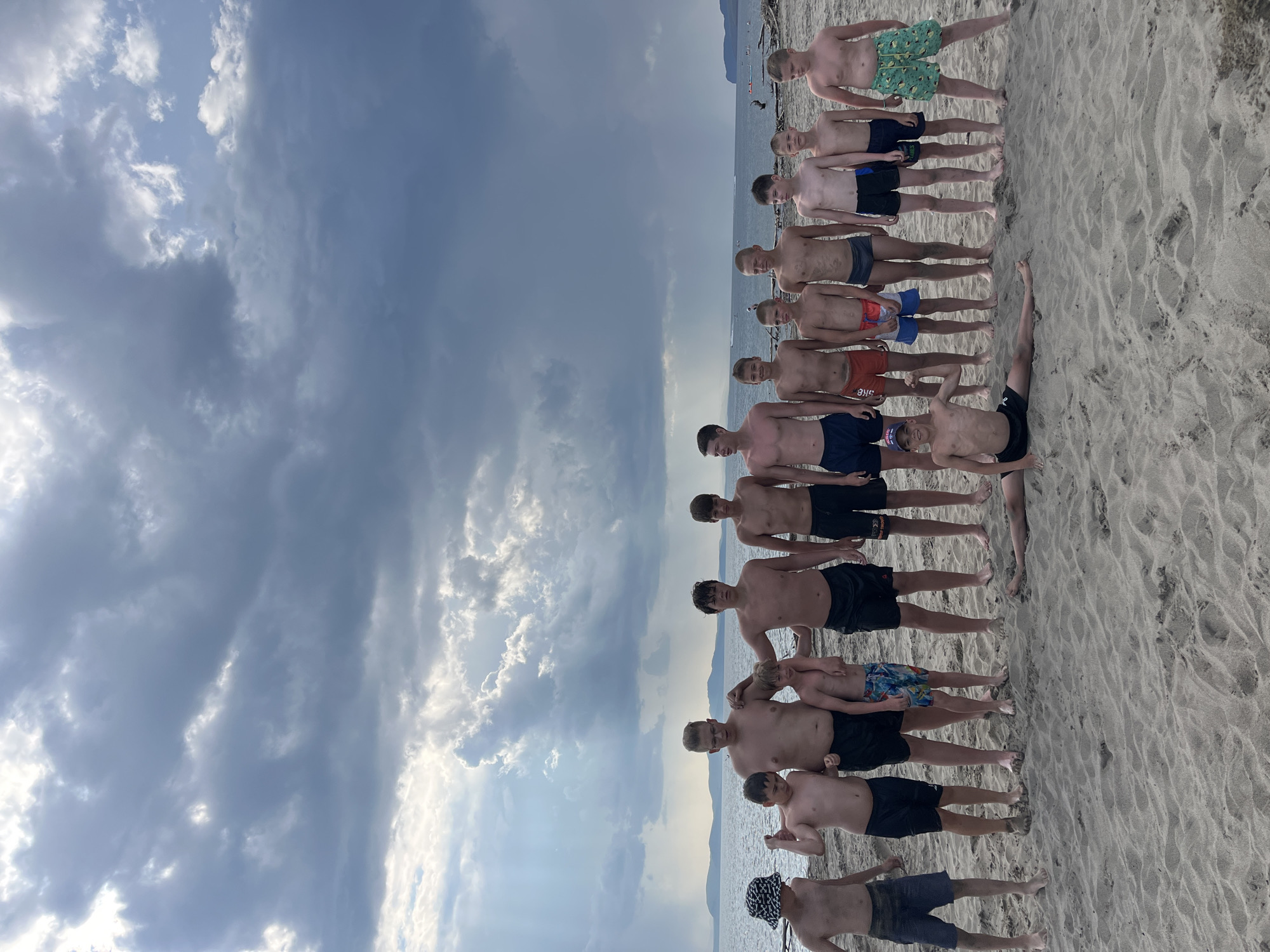 The image shows a group of boys on a sandy beach with the ocean and a cloudy sky in the background. The boys are standing in a line facing the camera, all wearing swim trunks. One boy in the foreground is doing a split on the sand. The sky is dramatic with dark clouds and shafts of light breaking through. The location is identified as the city of Krasnoturansk, Russia.