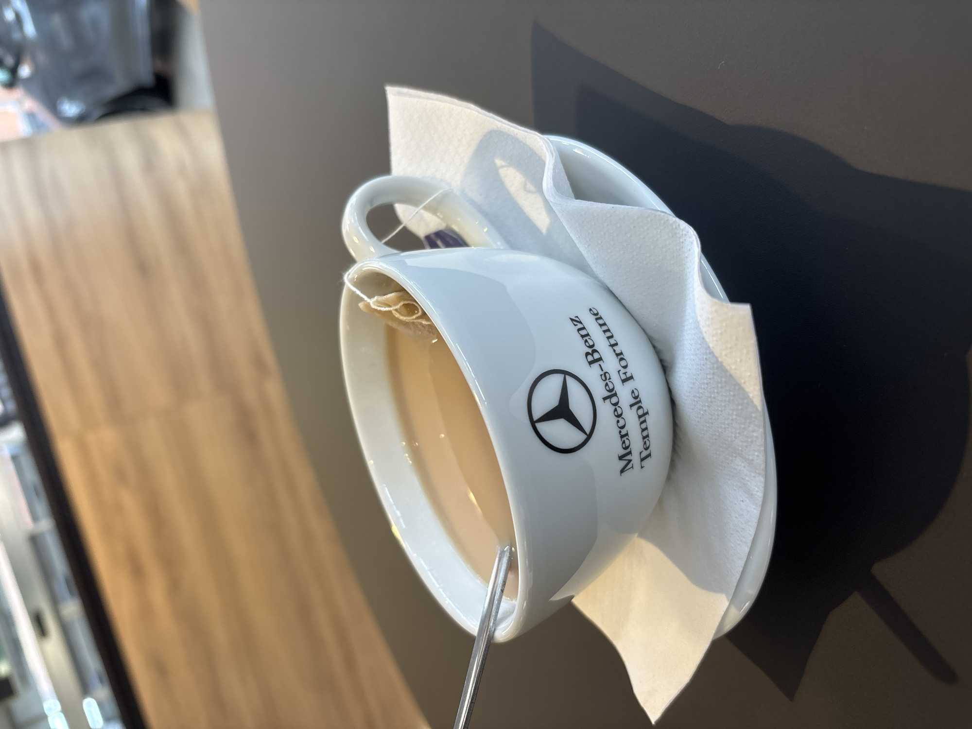 A teacup with a Mercedes-Benz logo sits on a table. The cup is white with the Mercedes-Benz emblem, along with the words "Mercedes-Benz Temple Fortune," printed on its side. A metal spoon is inside the cup, which appears to be filled with tea. A white napkin is wrapped around the cup. The table has a dark surface. The location is Temple Fortune in London, United Kingdom, as indicated by the text on the cup.