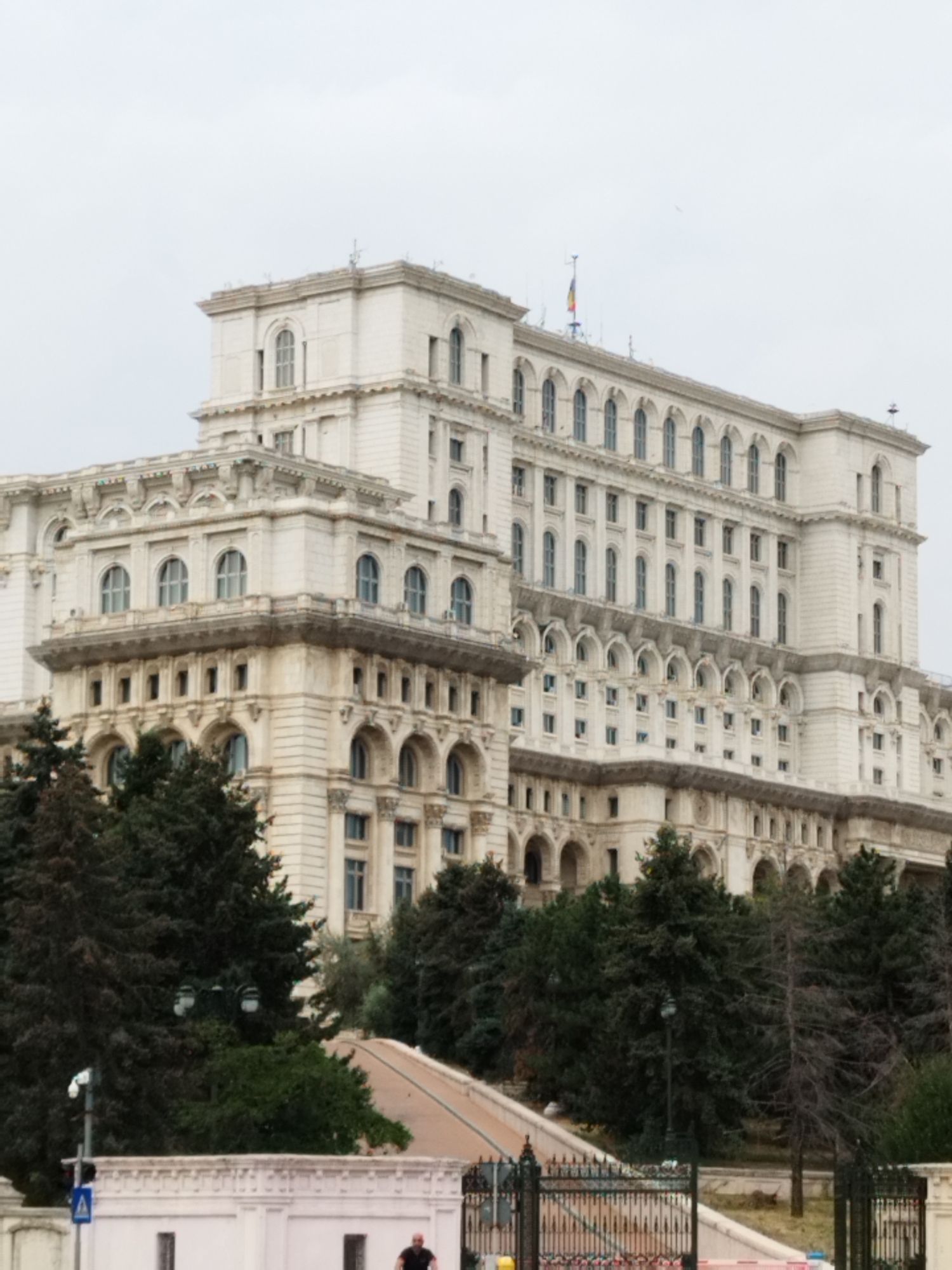This is a medium-shot, exterior, eye-level, vertical, full-color, daytime photograph of the Palace of the Parliament in Bucharest, Romania, on an overcast day. The immense, white, neoclassical building dominates the frame, its multiple stories featuring rows of arched windows and ornate architectural details. In the foreground, lush green trees partially obscure the lower levels of the structure. A paved ramp or walkway leads up towards the building from the lower right of the frame, and a black, ornate metal fence runs across the bottom portion of the image. In the far lower right, a person is visible riding a bicycle. The sky is a pale, uniform gray, indicating cloudy weather.