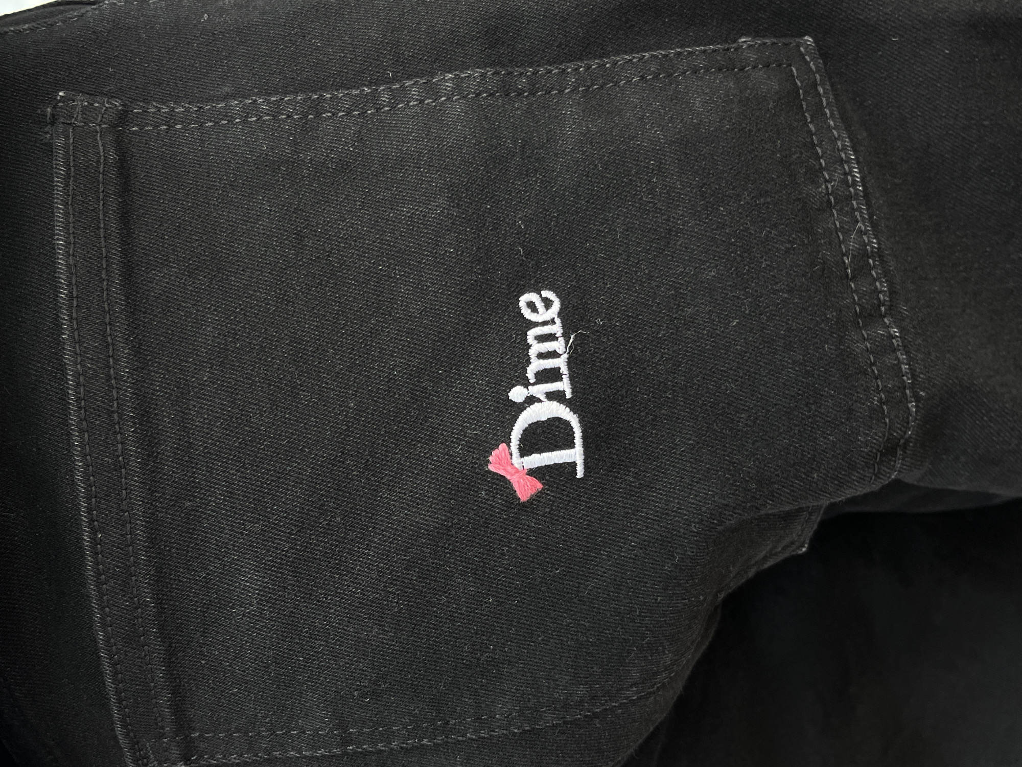 The image presents a close-up view of black denim fabric, likely from an article of clothing such as jeans or a jacket. The fabric exhibits a clear twill weave characteristic of denim. Light-colored stitching, appearing as white or very light grey, is visible outlining the edges of what appears to be a pocket or a structural panel on the garment. The stitching provides a strong contrast against the dark fabric.
Centered in the mid-right portion of the frame, a white embroidered logo spells out the word "Dime." The lettering is capitalized and features a distinct, slightly playful font. A notable detail is a small, vibrant pink bow embroidered directly to the left of the initial "D" in "Dime," adding a splash of color and a unique design element to the branding.
The image is focused on the texture of the fabric and the embroidered design, without any visible people or specific activities taking place. There are no explicit location cues, time of day indicators, or weather details within the image itself. The lighting is even, highlighting the fabric's texture and the embroidery's detail.