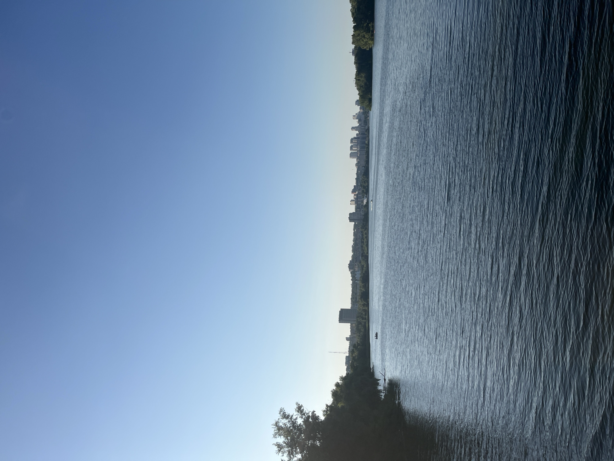 The image depicts a wide expanse of calm, rippling water, likely a river or lake, reflecting the clear blue sky. The water takes up the right half of the frame, with its textured surface creating a sense of depth and movement. Along the far bank, a cityscape is visible, characterized by numerous buildings, including a prominent tall structure. The buildings are nestled amongst lush green trees, suggesting a developed urban area on the waterfront. The far distance fades into a hazy horizon.
The left half of the image is dominated by a gradient of blue sky, transitioning from a deep azure at the top to a lighter hue towards the horizon. A solitary boat is a small detail in the water, close to the shore, and a few trees are visible in the foreground on the left, their dark silhouettes contrasting with the bright sky. The overall impression is one of tranquility and a bright, clear day, possibly late afternoon given the soft light. The scene suggests a peaceful moment captured in an urban setting that is embraced by nature. No visible text is present in the image.