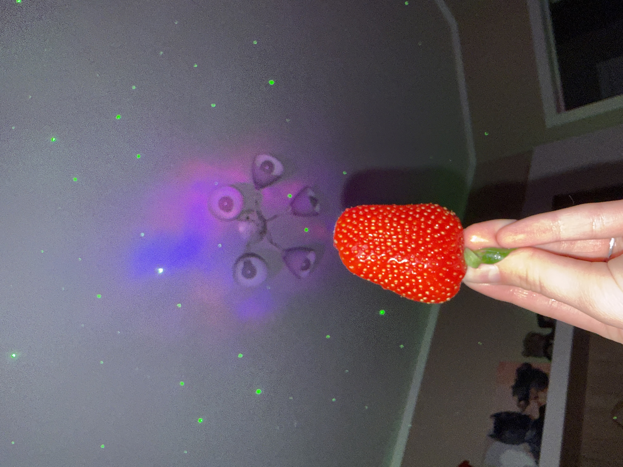 The image shows a hand holding a strawberry in front of a wall. The wall is decorated with green laser dots and a purple and blue light projection resembling eyes. The background is blurry and suggests an interior space. The location appears to be indoors.