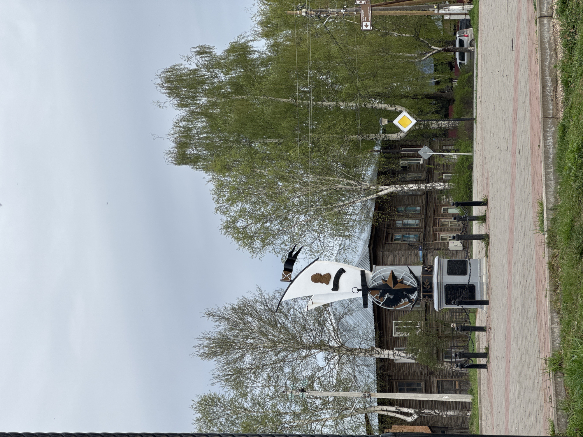 The image shows a street scene in the city of Osa, Russia. A monument featuring a stylized ship sail stands in the foreground. The sail is white with a black design on it. Behind the monument is a traditional wooden building with multiple windows. A road with a sidewalk runs along the right side of the image. There are birch trees lining the street, and utility poles with wires are visible above them. A yellow diamond-shaped road sign is also present. The sky is overcast.