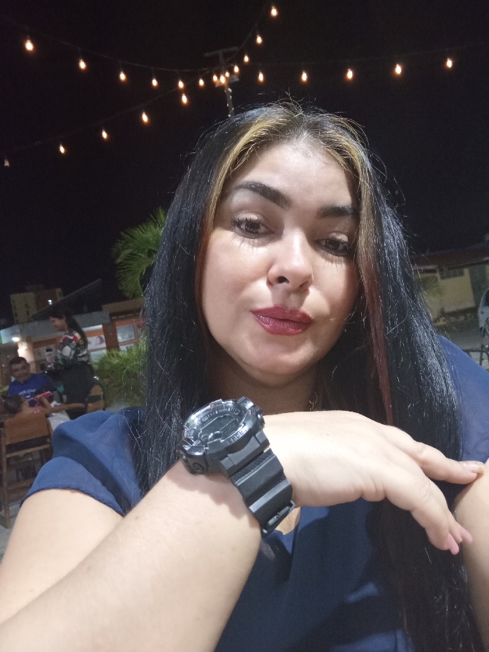A woman poses at night under string lights. She has dark hair with blonde and red highlights, and is wearing dark lipstick. She wears a large, black digital watch on her left wrist. Other people are visible in the background, including a woman in a floral shirt and a man in a blue shirt seated at a table. Green foliage and buildings are also in the background.