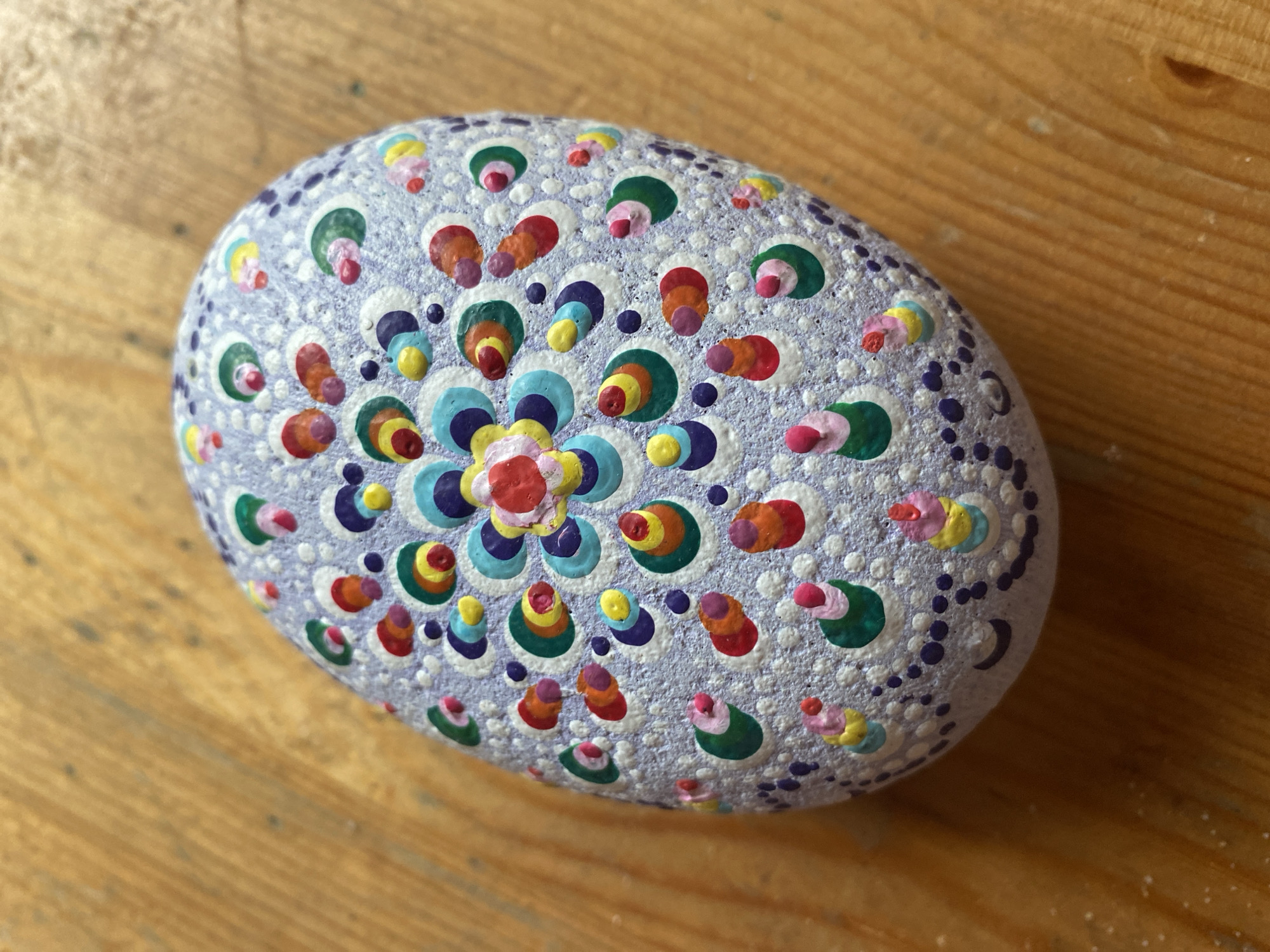 A purple, oval-shaped stone sits on a wooden surface. The stone is decorated with a mandala pattern created with small dots and circles of various colors, including white, purple, blue, green, yellow, orange, and red. The mandala features a central floral design with concentric rings of color, surrounded by radiating patterns of dots and colorful circular motifs. The wooden surface appears to be a table or countertop with visible grain and texture. The image is taken from a top-down perspective.