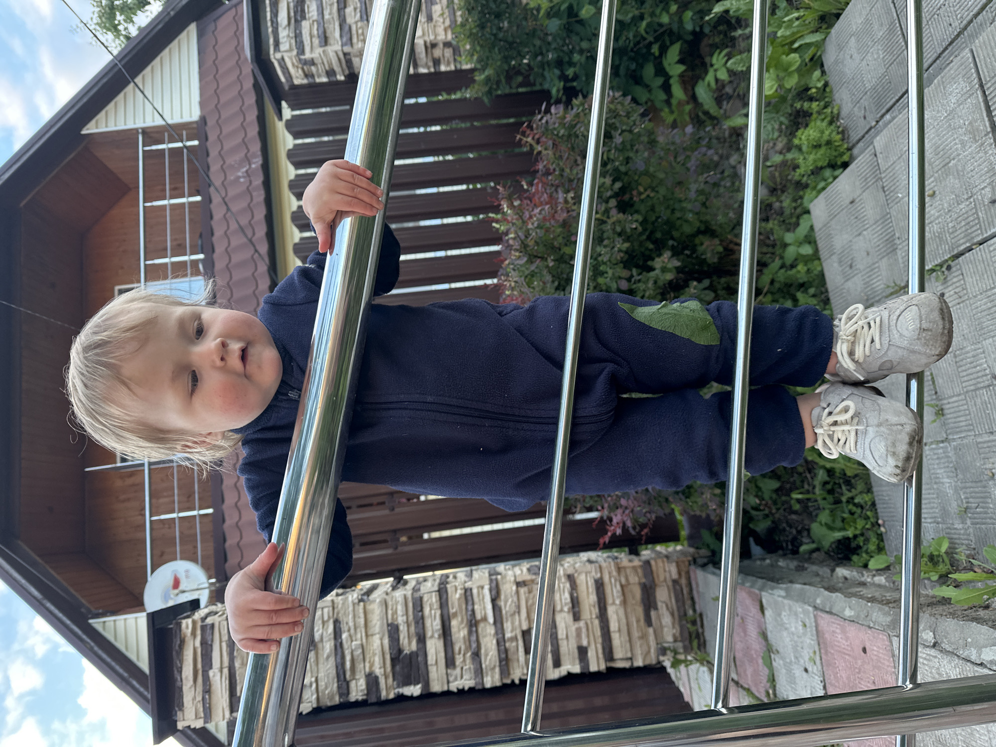 The image shows a young child, likely a toddler, leaning on a metal railing. The child has fair hair and is wearing a dark blue fleece jumpsuit and light-colored sneakers. The child is looking towards the camera with a neutral expression.
The setting appears to be outdoors, possibly a backyard or a porch, with a house visible in the background. The house has a brown roof and siding, and a satellite dish is mounted on the wall. There is lush greenery surrounding the area, with trees and bushes. The ground is paved with rectangular tiles. The lighting suggests it is daytime, possibly late afternoon or early evening, as the sunlight creates some shadows.
The child is holding onto the railing with one hand, and their feet are also visible, supported by the railing and the ground. The overall impression is a candid snapshot of a child exploring their surroundings. There is no visible text in the image. The location context provided is Nizhny Novgorod, Russia.
