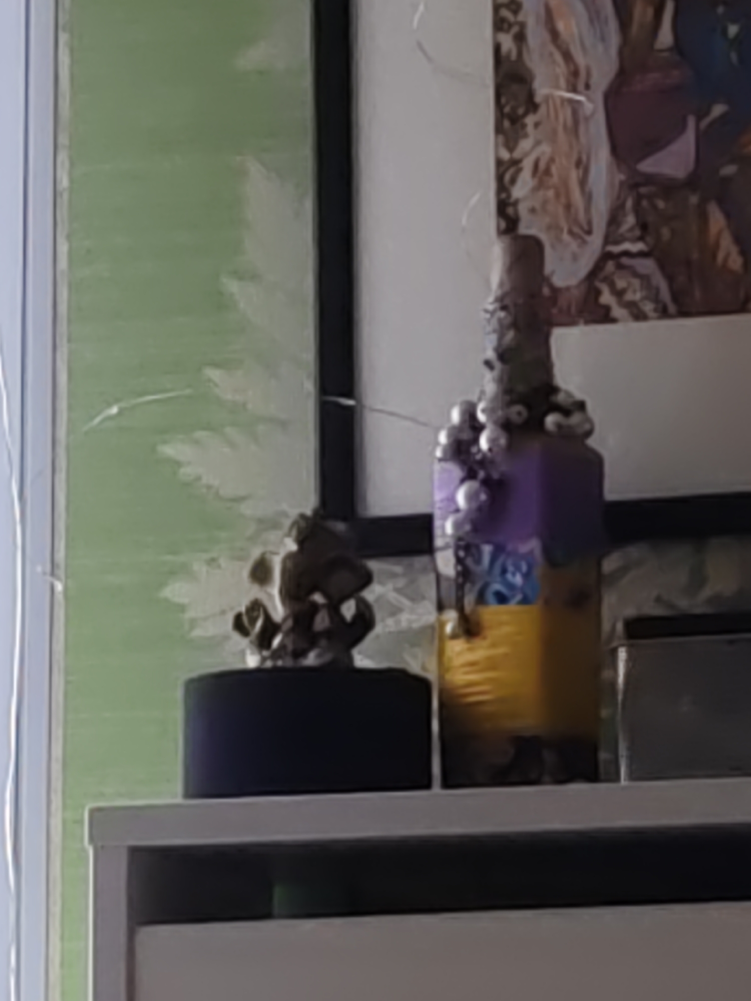 A close-up, slightly out-of-focus shot shows a decorative bottle with purple and yellow sections, adorned with silver spheres, standing on a white shelf. To the left of the bottle, a small, dark object with an intricate, possibly metallic, design sits on a black cylindrical base. The background features a green wall with a faint, light-colored pattern and a framed image with abstract colorful shapes. The scene is indoors, likely in a residential setting, with no discernible details indicating a specific location within Rostov-on-Don, Russia.