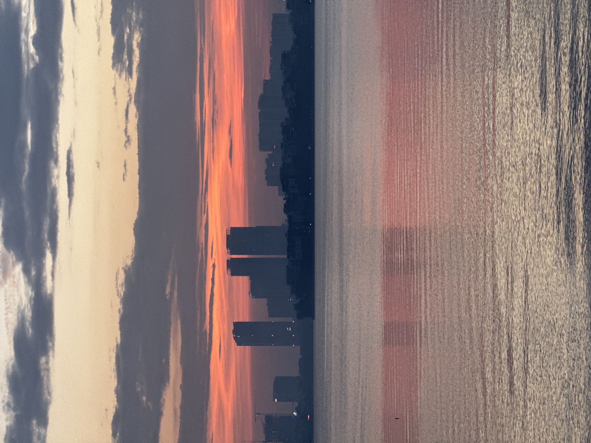 This is a photograph of the skyline of Hanoi, Vietnam, at sunset. The image is dominated by the vibrant colors of the sky, with hues of orange, pink, and purple blending together. Dark clouds are scattered across the sky, creating a dramatic contrast with the colorful sunset. In the distance, the silhouettes of tall buildings and structures can be seen against the sky, indicating an urban environment. A large body of water, likely a lake or river, occupies the foreground of the image, with its surface reflecting the colors of the sky and the city lights. The water is calm, with subtle ripples creating a textured effect. The overall scene depicts a serene and picturesque urban landscape at dusk.