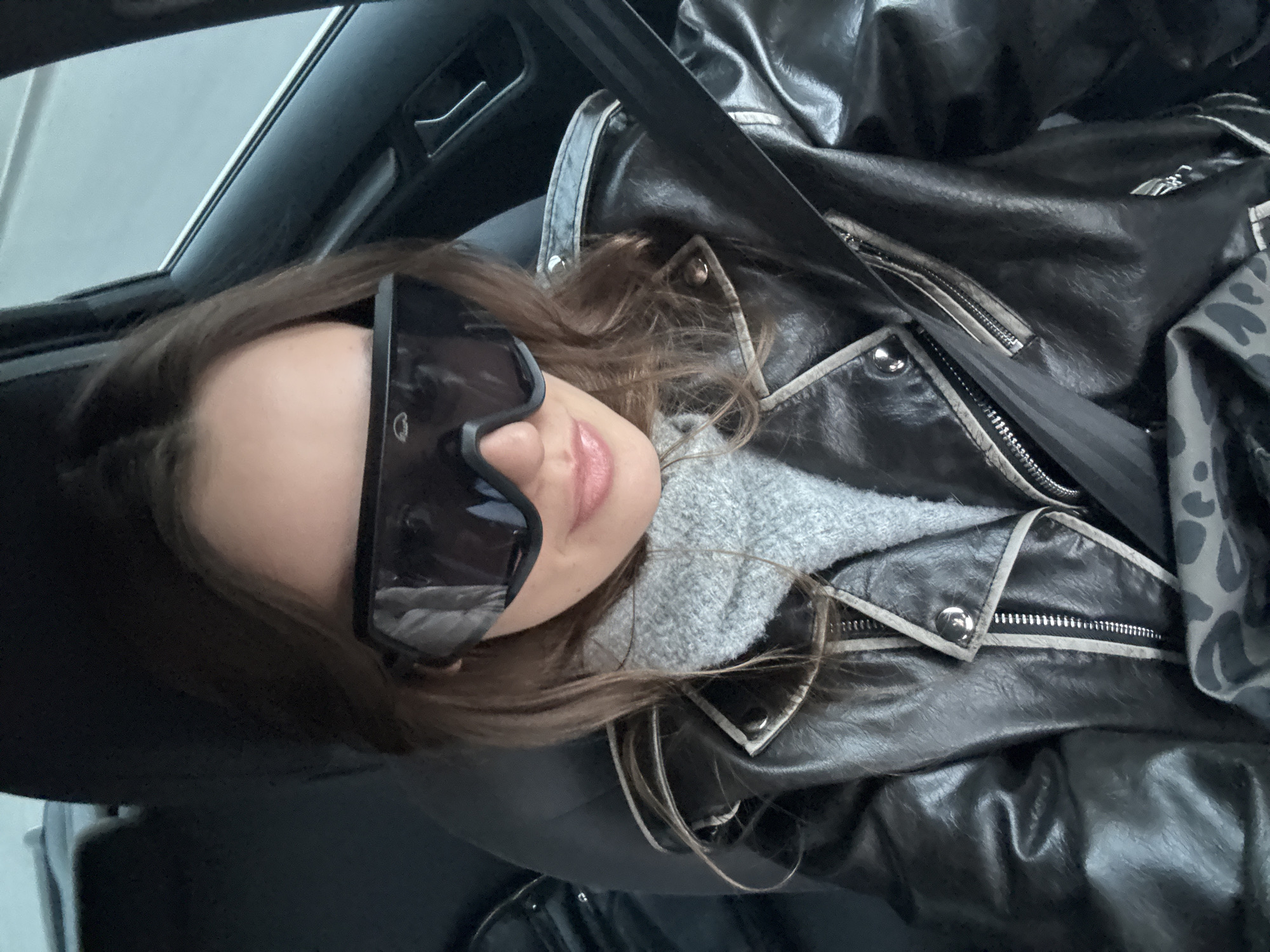 A woman is in a car, wearing oversized sunglasses and a black leather jacket with a grey sweater underneath. She has brown hair and is wearing a seatbelt. The location is unknown, and there is no evidence to suggest it is in Krasnoyarsk, Russia.