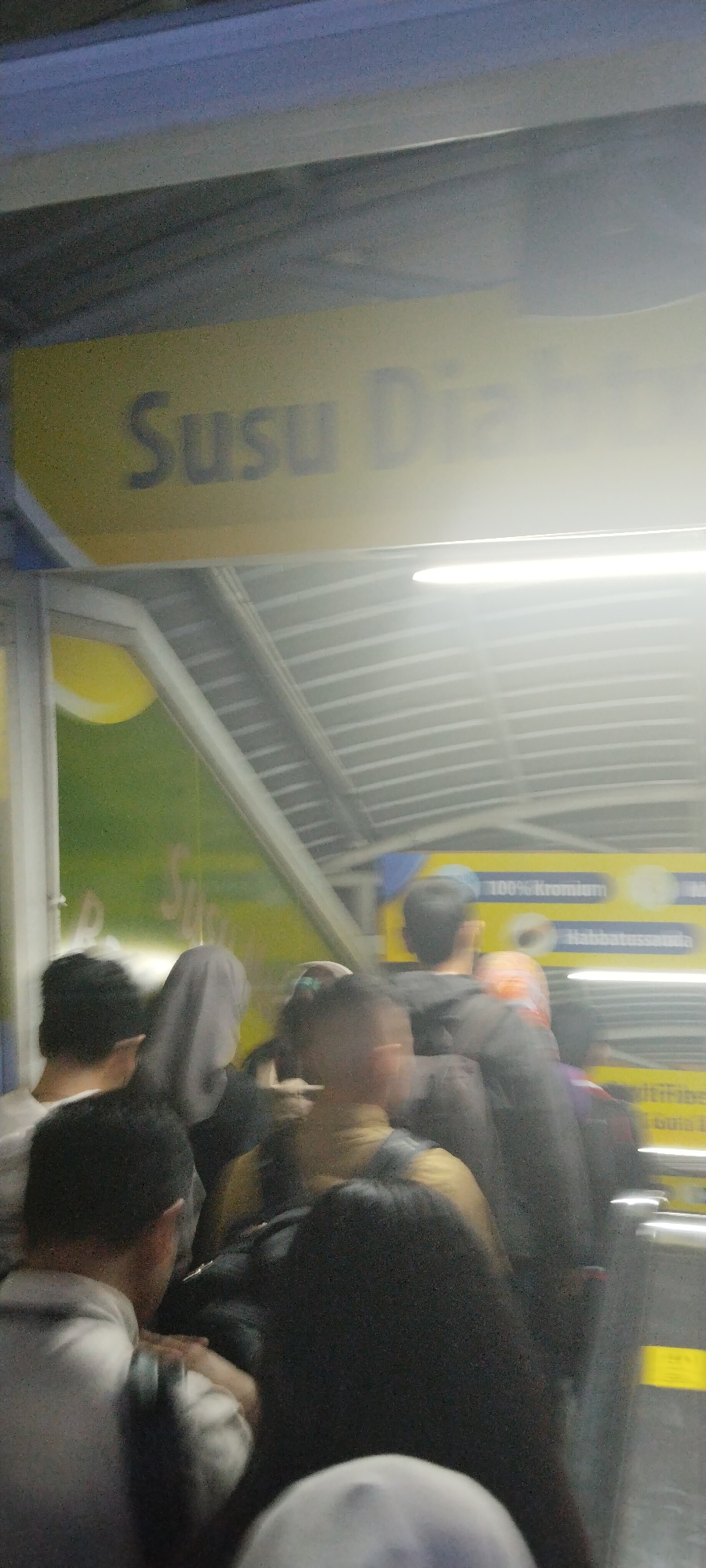 The image captures a blurry view of a crowded area, possibly a public transportation hub or a busy walkway in Central Jakarta, Indonesia. A yellow sign with dark text, partially obscured and out of focus, is visible at the top, with the legible portion reading "Susu Dia". Below this sign, a silver-colored corrugated roof structure is evident, illuminated by bright lights. A group of people, also out of focus and appearing to be in motion, are seen in the foreground and midground. Their faces and specific features are indistinct due to the blurriness. The overall impression is one of a bustling, active environment with movement and a crowd. The lighting suggests it might be indoors or under a covered area, possibly with artificial lighting contributing to the overall brightness and haziness.