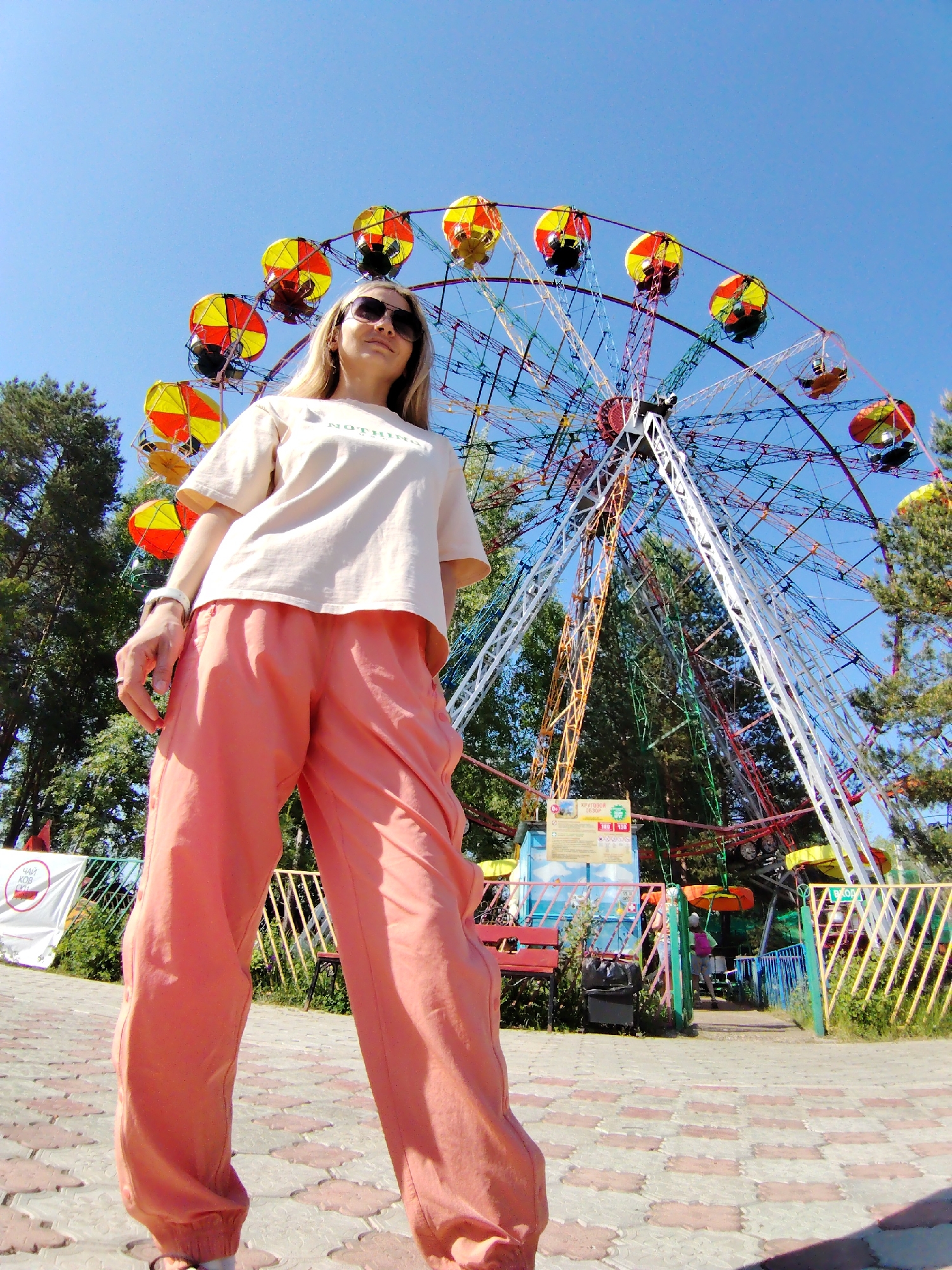 The image captures a full-body shot of a woman standing in the foreground, with a colorful Ferris wheel prominently featured in the background. The woman wears sunglasses, a light-colored t-shirt, and salmon-colored pants.
The Ferris wheel has a multicolored structure and cars painted in yellow and orange. Trees surround the Ferris wheel, suggesting a park setting. The ground is paved with patterned bricks.
The location is in the city of Chaikovsky, Russia, identified by signage visible in the scene. Additional details such as benches and fencing can also be observed.