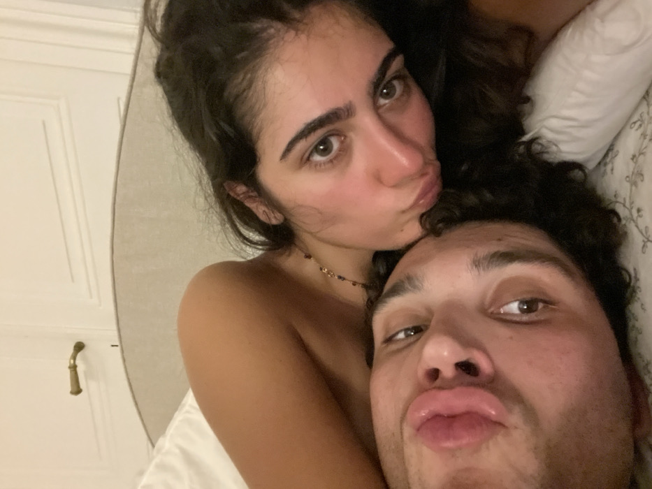 A selfie features a couple lying in bed. The woman, with dark hair and fair skin, is above the man. Both are puckering their lips. The woman wears a delicate necklace. Behind them are pillows and bedding with a subtle floral pattern. To the left, a glimpse of a white door with a gold handle is visible. The lighting is soft and warm.