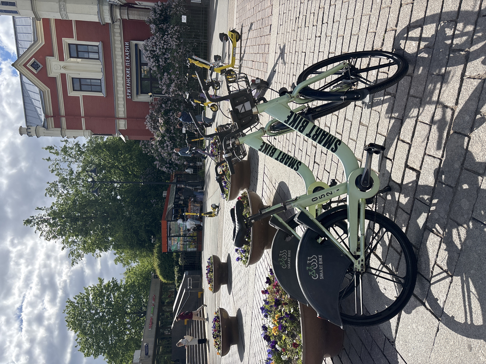 The image shows a row of Smart Bikes parked on a cobblestone path in Saint Petersburg, Russia. A red brick building with signage reading "БРИТАНСКИЕ ПЕКАРНИ" (British Bakery) is visible in the background. Several Smart Bikes, recognizable by their light green frames and "SMART BIKE" lettering, are lined up near flower planters. Some scooters are also visible in the background. People are seen in the background. The sky is cloudy. A metro station sign is visible on the left side of the frame, indicating a public transportation hub.