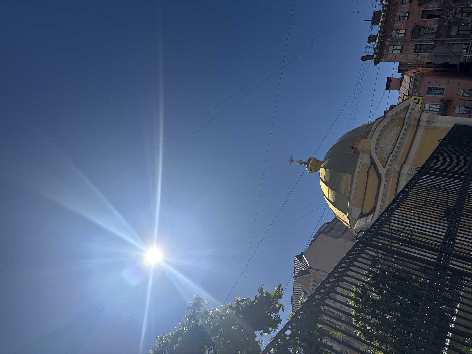 The image captures a bright, sunny day in Saint Petersburg, Russia, with a clear blue sky dominating the upper half. The sun is prominently visible in the upper left quadrant, emitting strong rays and lens flare. In the foreground, a black metal fence with decorative circular patterns stretches across the lower portion of the image. Behind the fence, the golden dome of a church, topped with a cross, is partially visible. The dome is a striking yellow-gold color. To the right of the dome, older buildings with red and pink facades, featuring multiple windows, are visible. Some of these buildings appear to be residential. Overhead, several power or utility lines crisscross the sky. To the left of the dome and fence, the lush green leaves of a tree are visible, adding a touch of nature to the urban scene. The overall impression is a view of an historic religious building and surrounding architecture on a clear, sunny day.
