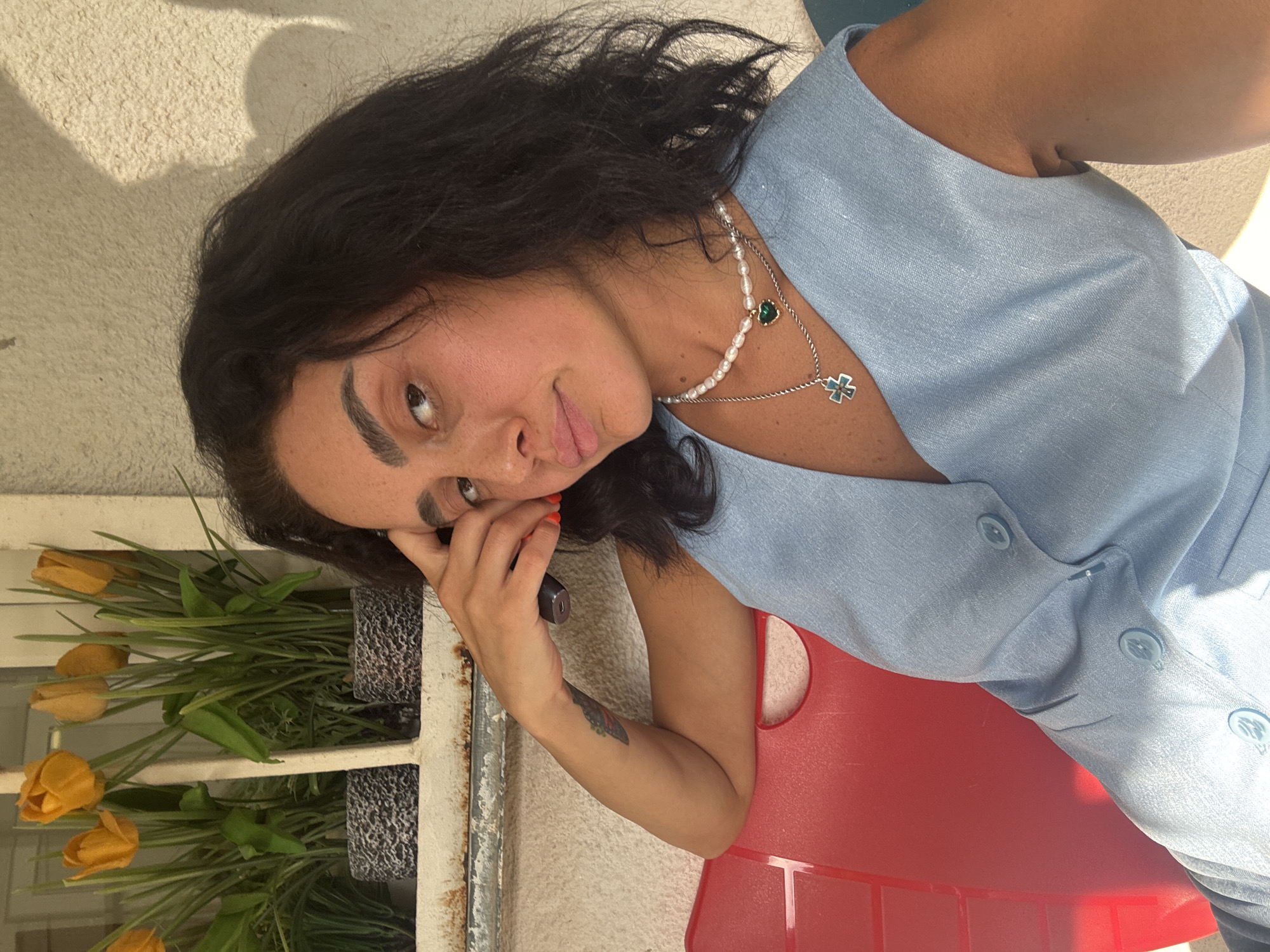 The image shows a woman sitting on a red chair outdoors. She has dark, wavy hair and is wearing a light blue sleeveless top with buttons down the front. She is also wearing a pearl necklace with a green heart pendant and a silver chain with a cross pendant. She is holding a dark object, possibly a phone, up to her face with her right hand. Her left arm rests on her lap, and a tattoo is visible on her forearm. In the background, there are potted plants with orange tulips on a windowsill. The wall behind her is textured and light-colored, with a shadow cast on it. The overall setting appears to be an outdoor area in a city, with the context of Tbilisi, Georgia, suggesting it might be a residential or public space within the city.
