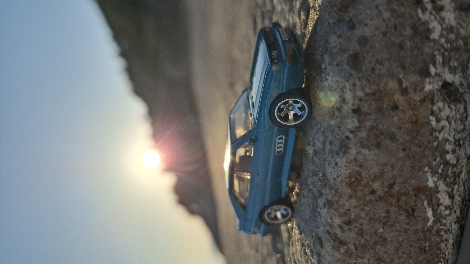 A turquoise toy car with the Audi logo is positioned on a gray, rough surface that resembles stone or rock, under bright sunlight that casts lens flare. The background shows a blurry mix of light blue sky and possibly a hill or cliff. The location is likely Boreti, Montenegro, given that the image has been specified to be there.