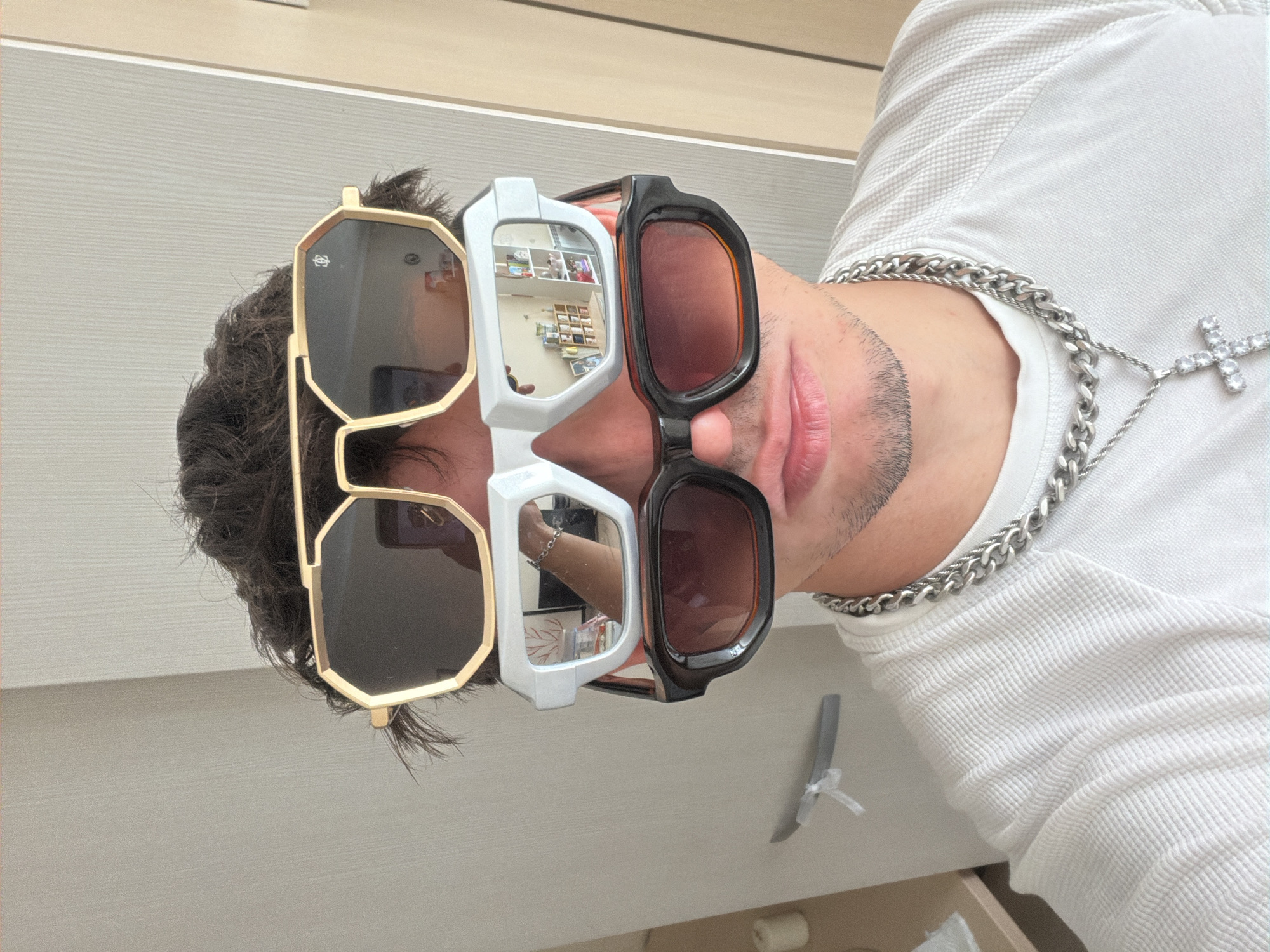 A man is pictured wearing three pairs of sunglasses, a silver chain, and a white waffle-knit shirt. The sunglasses are a gold, a white, and a dark brown pair. The man has short, dark hair and a neatly trimmed beard. The background features a light-colored wall and a section of a drawer or shelf. The location is a residential space, presumably in the city of Sofia, Bulgaria.