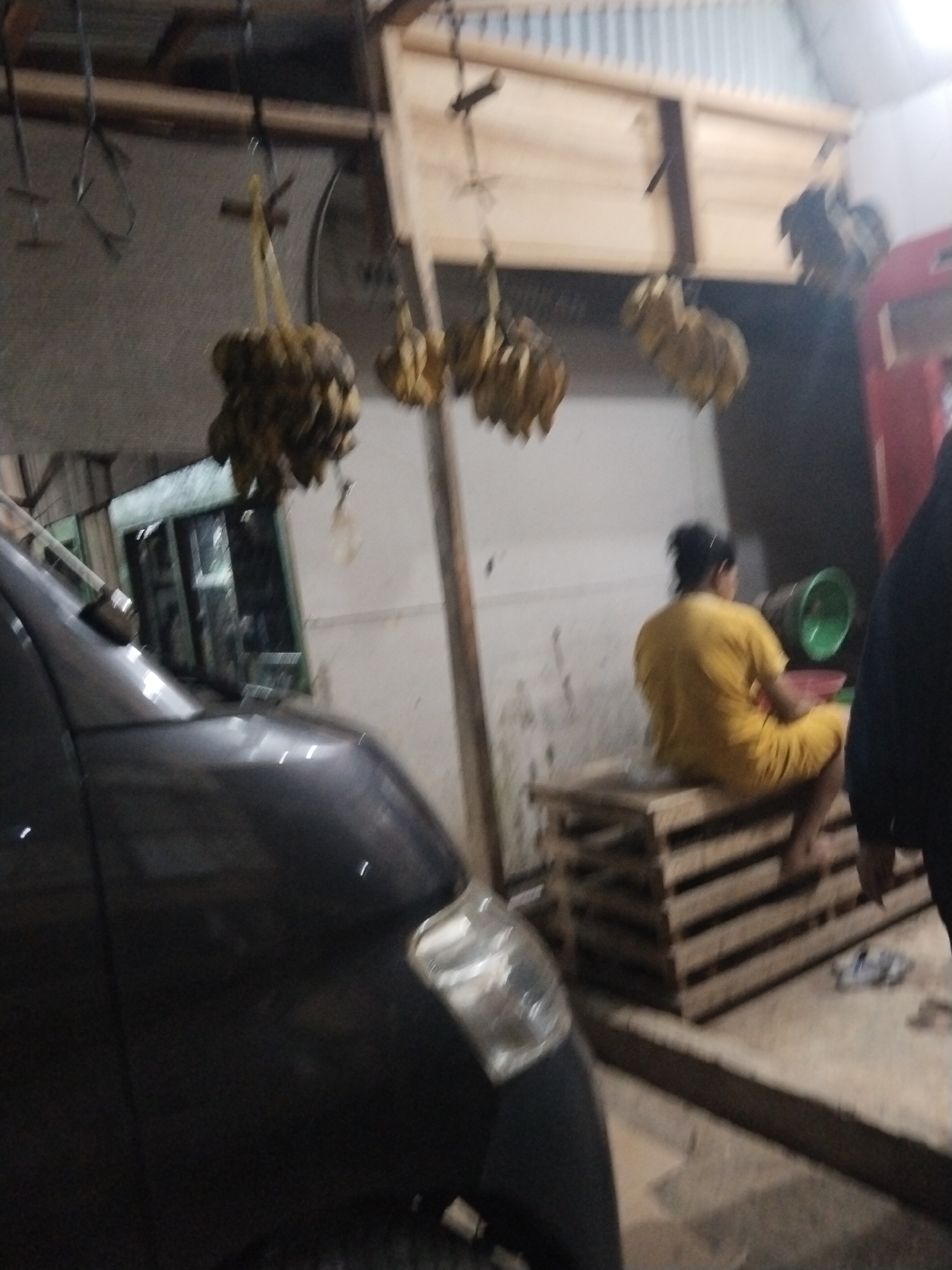 The image depicts an indoor scene, likely a market or storage area, in Suka Ramai, Indonesia. In the foreground, the dark, reflective surface of a vehicle is visible, possibly a car or van. In the background, a woman wearing a yellow outfit is seated on a stack of wooden pallets. To her right, a green fan and a red bowl are present. Above, bunches of what appear to be dried bananas or similar produce are hanging from the ceiling. The environment appears utilitarian, with corrugated metal roofing and concrete or dirt flooring. The lighting suggests it is either early morning or late evening.