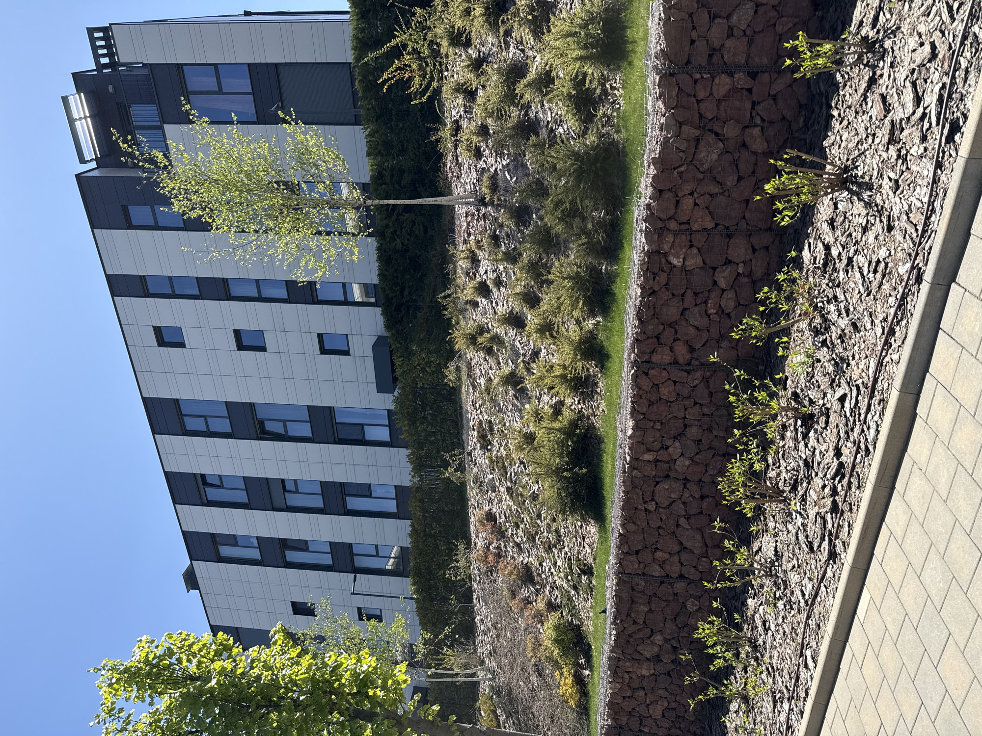 The image depicts a modern architectural scene, possibly in Sofia, Bulgaria. A multi-story building with a black and white facade is visible on the left. A terraced landscaping feature occupies the right side of the frame, consisting of tiered sections with vegetation and retaining walls constructed from rocks held within wire mesh. The bottom tier has bark mulch with small shrubs. A paved walkway is situated to the right of the landscaping. The sky is clear and blue. The image is oriented vertically.