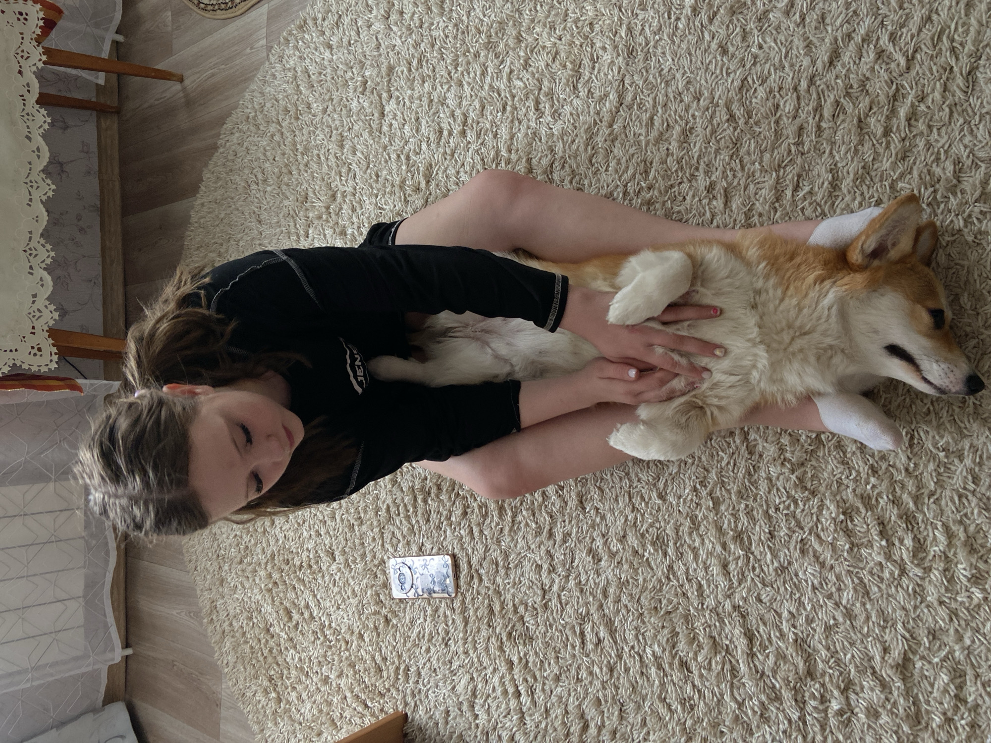A young girl in a black long-sleeved shirt lies on a beige carpet, cradling a corgi dog on its back. A small, rectangular object with a patterned surface rests near her legs on the carpet. The background shows parts of a wooden chair and a white lace-edged tablecloth, indicating an indoor setting.
The photo was taken in Chaikovsky, Russia.