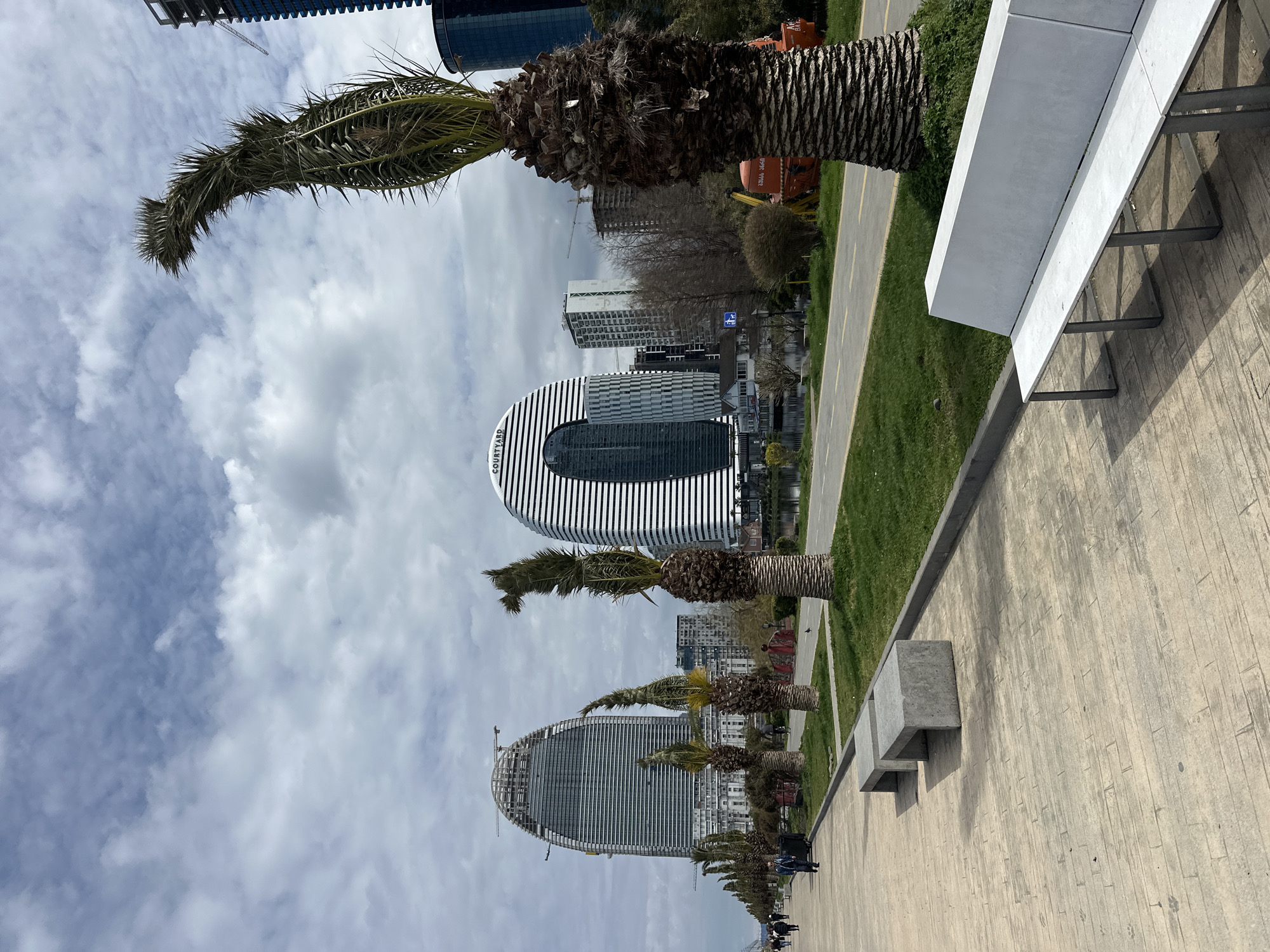 The image shows a promenade in Batumi, Georgia, lined with palm trees and benches. The architecture is modern, with the Courtyard by Marriott Batumi hotel visible, along with other high-rise buildings, including one under construction. The sky is cloudy. People are walking along the promenade.