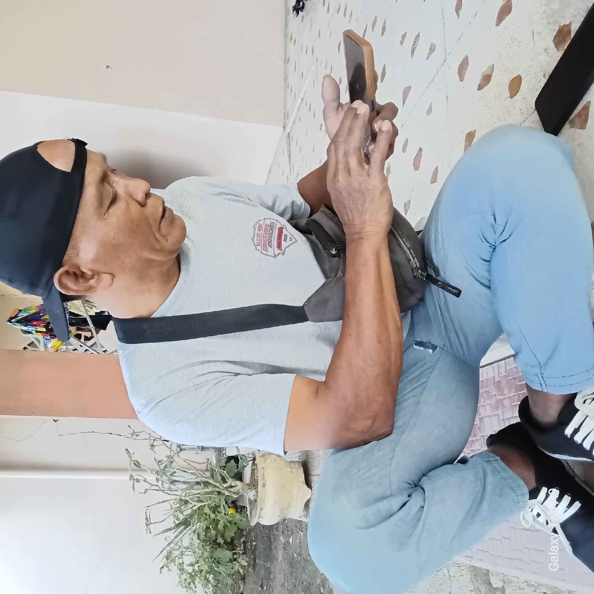 The image depicts a man squatting on a tiled patio outside a building in Sambirejo, Indonesia. He is wearing a light grey t-shirt with a logo that appears to read "BULENG SPANDI CROSSMAX MOTOCROSS 2023", light blue pants, a black baseball cap, and black and white sneakers. He has a dark grey crossbody bag slung over his shoulder and is holding a brown-cased smartphone in his hands, appearing to interact with it. Another black smartphone is visible on the ground to his right. In the background, there is a white wall with a crack, a potted plant with dry branches, and a white column. The floor is made of light-colored tiles with scattered brown oval-shaped patterns. The overall lighting suggests it is daytime. The text "Galaxy Tab" is partially visible in the bottom left corner of the image.