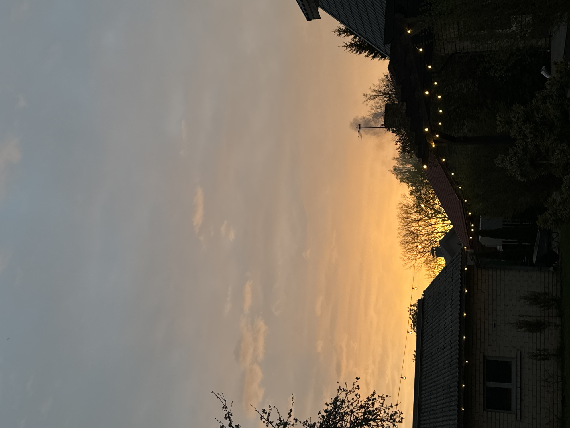 The image shows a view in Carnikava, Latvia, at sunset or sunrise. The sky is a gradient of gray to orange. Several buildings are visible, including a brick house with a tiled roof and lit with string lights. Another building with a dark roof also has string lights. Trees are silhouetted against the sky. There is an antenna on top of one of the buildings. The scene appears calm and quiet.