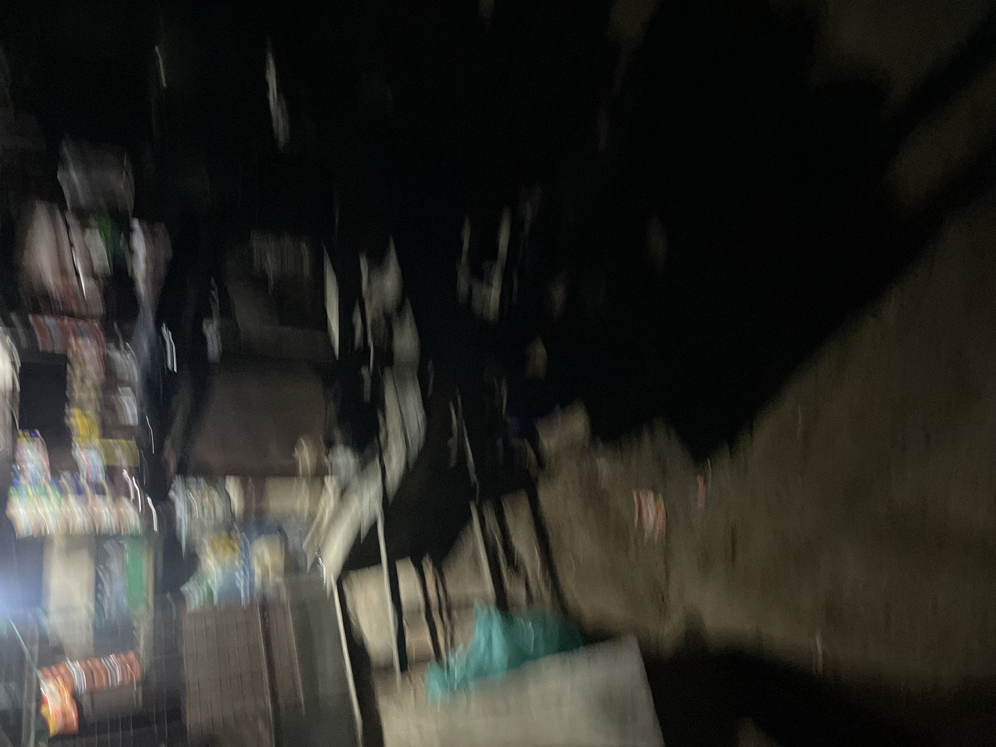 The image depicts a blurry, low-light scene that appears to be an outdoor market stall. In the background, there are shelves stocked with various items, possibly packaged goods, with some colorful wrappers visible. A teal-colored plastic bag is hanging to the left, and a large dark shadow dominates the right side of the frame. The ground in the foreground is a dirt or asphalt surface, with some debris scattered on it. The overall impression is of a dimly lit, possibly nocturnal, commercial setting in what could be Safari, Nigeria.