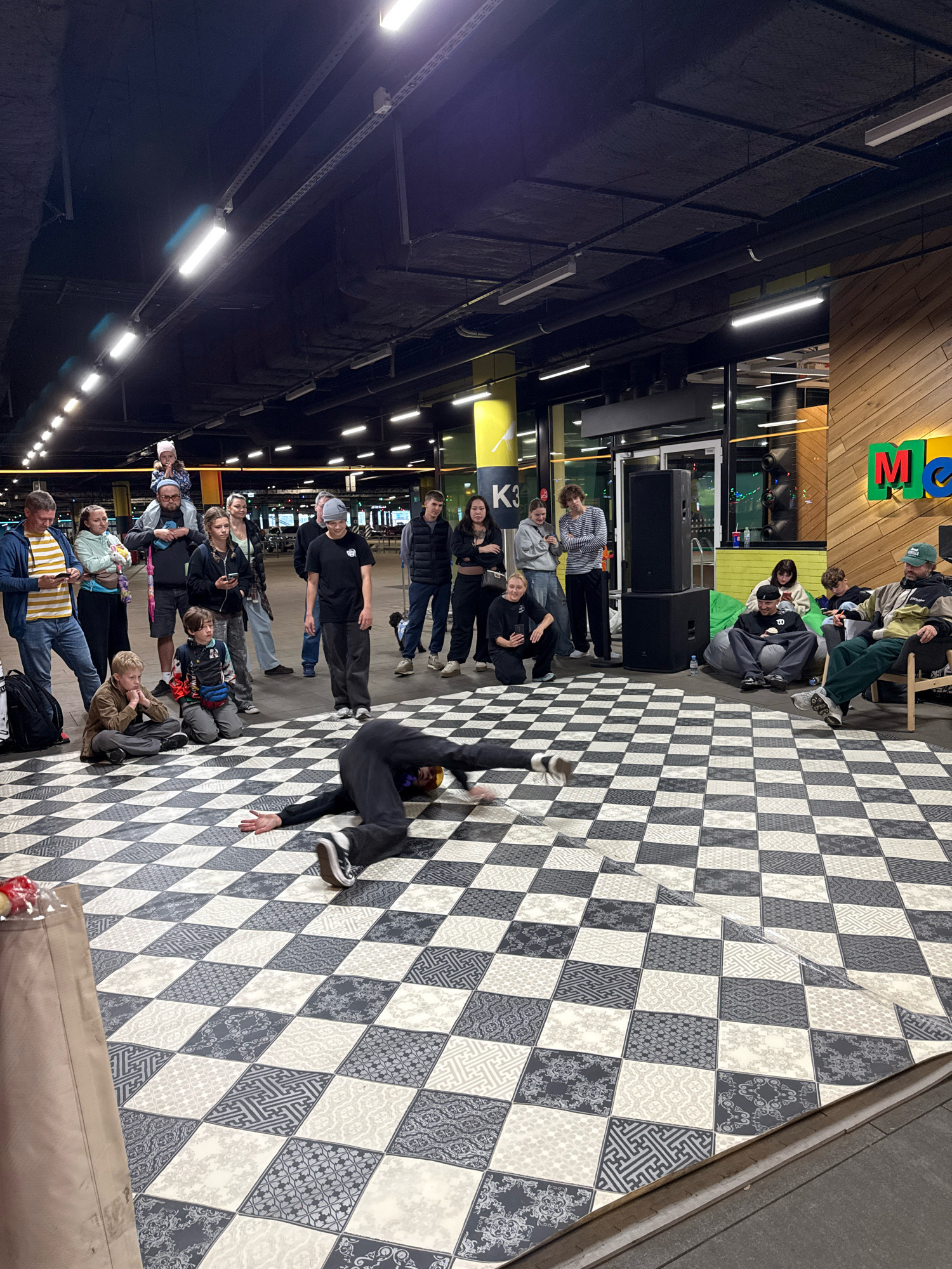 An outdoor performance is taking place on a checkered floor in Saint Petersburg, Russia. A breakdancer in black clothing is in mid-move, with one leg extended upwards and the other bent. A crowd of spectators, consisting of adults and children, are watching the performance. Some individuals are standing, while others are seated. Behind the crowd, there are building structures with large windows and neon signage. The environment appears to be a public space, possibly a plaza or a street. The lighting is provided by overhead fluorescent lights and some ambient natural light.
