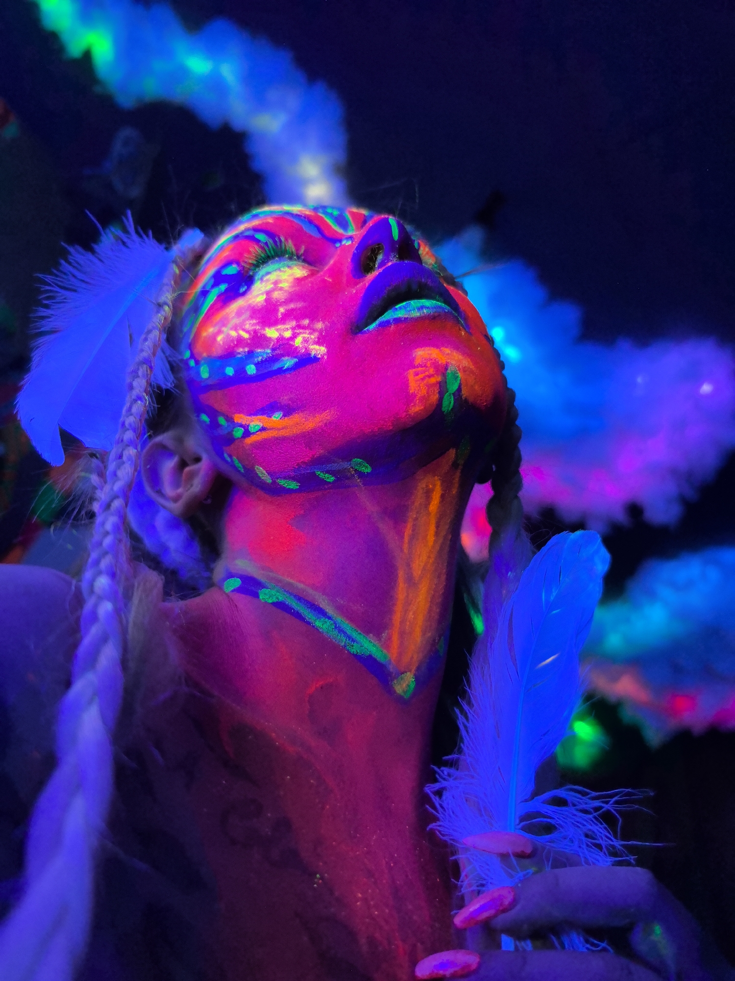 The image captures a person, likely female, illuminated by UV light in a dark setting, possibly indoors. The subject's face, neck, and upper body are elaborately painted with vibrant, neon-colored, glow-in-the-dark makeup in intricate patterns of pink, green, blue, and orange. Their eyes are closed, and their head is tilted upwards, creating a contemplative or ethereal pose.
Two white feathers are visible: one tucked behind the left ear, blending with the person's light-colored braided hair, and another held delicately in the right hand. The nails on the visible hand are painted with bright pink polish, also glowing under the UV light.
The background features blurred, glowing forms resembling clouds or smoke, rendered in shades of bright blue, purple, and hints of green, suggesting an atmospheric or mystical ambiance. The overall impression is one of a vibrant, artistic performance, a party, or a photoshoot utilizing blacklight effects. The intense luminescence of the body paint and accessories contrasts sharply with the dark surroundings.
There is no discernible text visible in the image. The scene is characterized by its powerful use of color and light, creating a surreal and energetic visual experience.