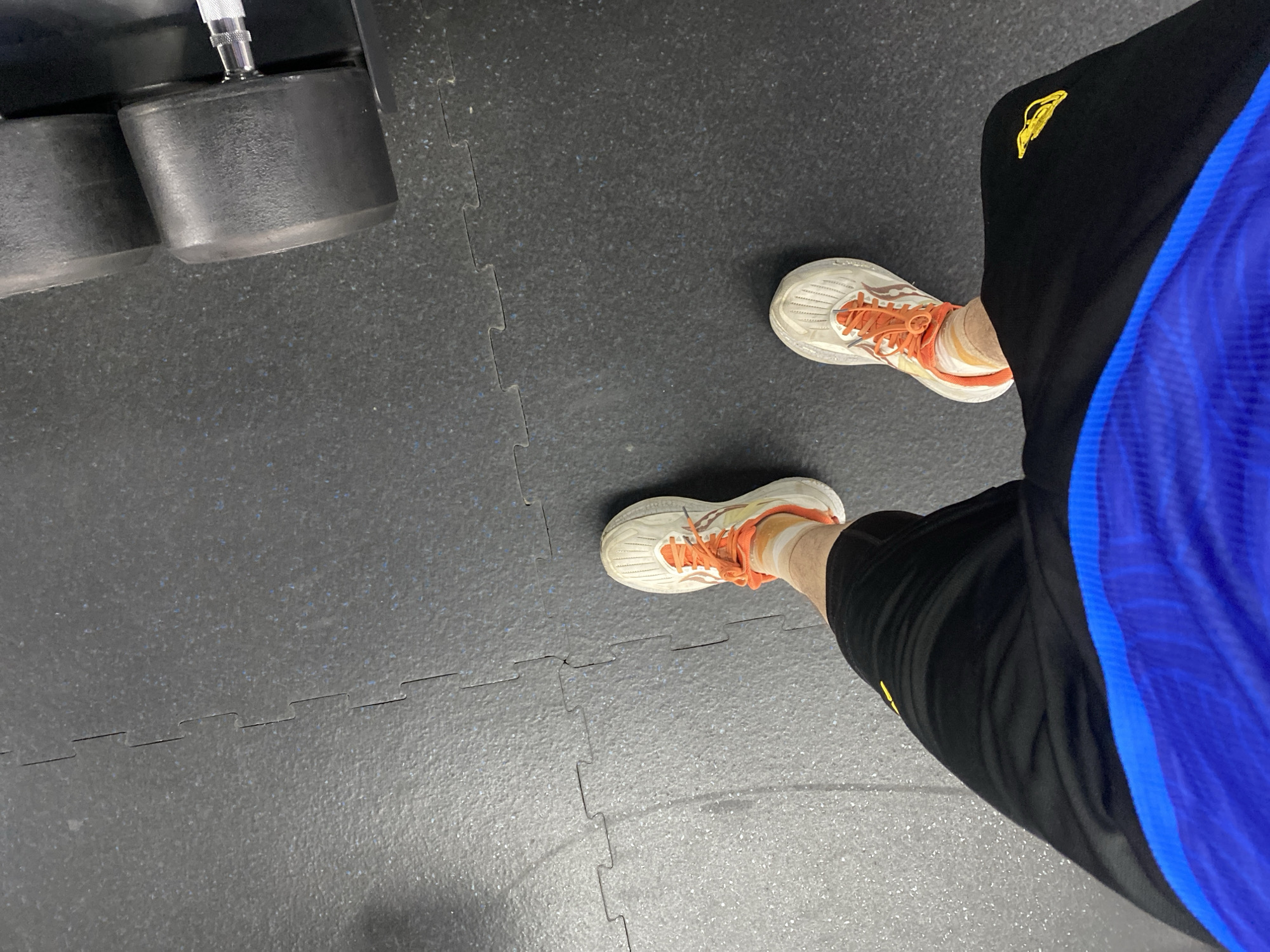 The image shows a pair of feet wearing athletic shoes with orange laces and black shorts with blue accents, standing on a black gym floor made of interlocking mats. A dumbbell is visible in the upper right corner of the frame. The scene is indoors and appears to be in a gym. There is no indication of the city of Astana, Kazakhstan in the image.