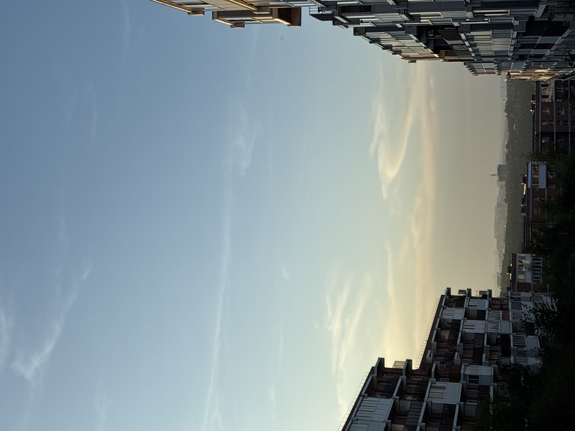 In Markova, Russia, a dusk sky stretches over a cityscape. The scene is dominated by the facades of apartment buildings. On the left, a multi-story building with a red-brick base and white balconies is silhouetted against the fading light. To the right, another apartment building, taller and with a gray facade and dark balconies, rises sharply into the frame. Between these buildings, a hazy horizon reveals a distant urban landscape dotted with trees and structures, including a prominent tall building with a crane. The sky itself is a gradient of pale blue to a warm, muted yellow near the horizon, with wispy clouds catching the last rays of sunlight. No people or discernible actions are visible, creating a still, architectural portrait of the city at twilight.