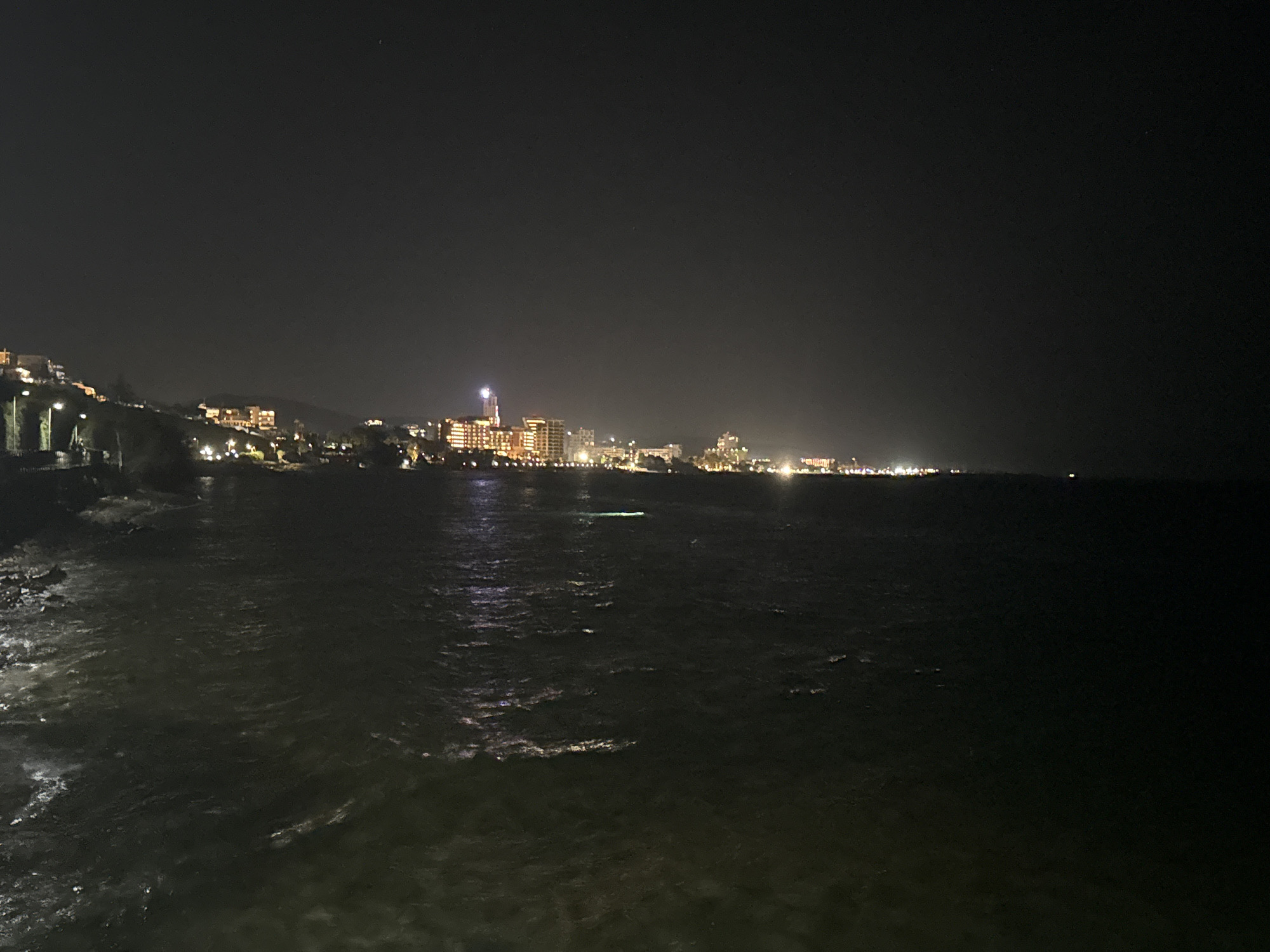 The image captures a night scene of a coastal city, likely within the Agios Tychona Community in Cyprus. A body of dark water, reflecting the lights of the city, occupies the foreground. In the background, a sprawling city skyline is illuminated by numerous lights from buildings and street lamps. Some structures are taller than others, suggesting a mix of residential and commercial buildings. The lights create a warm glow against the dark sky. The scene appears tranquil, with no visible people or boats. The overall impression is one of a well-lit city by the sea at night.