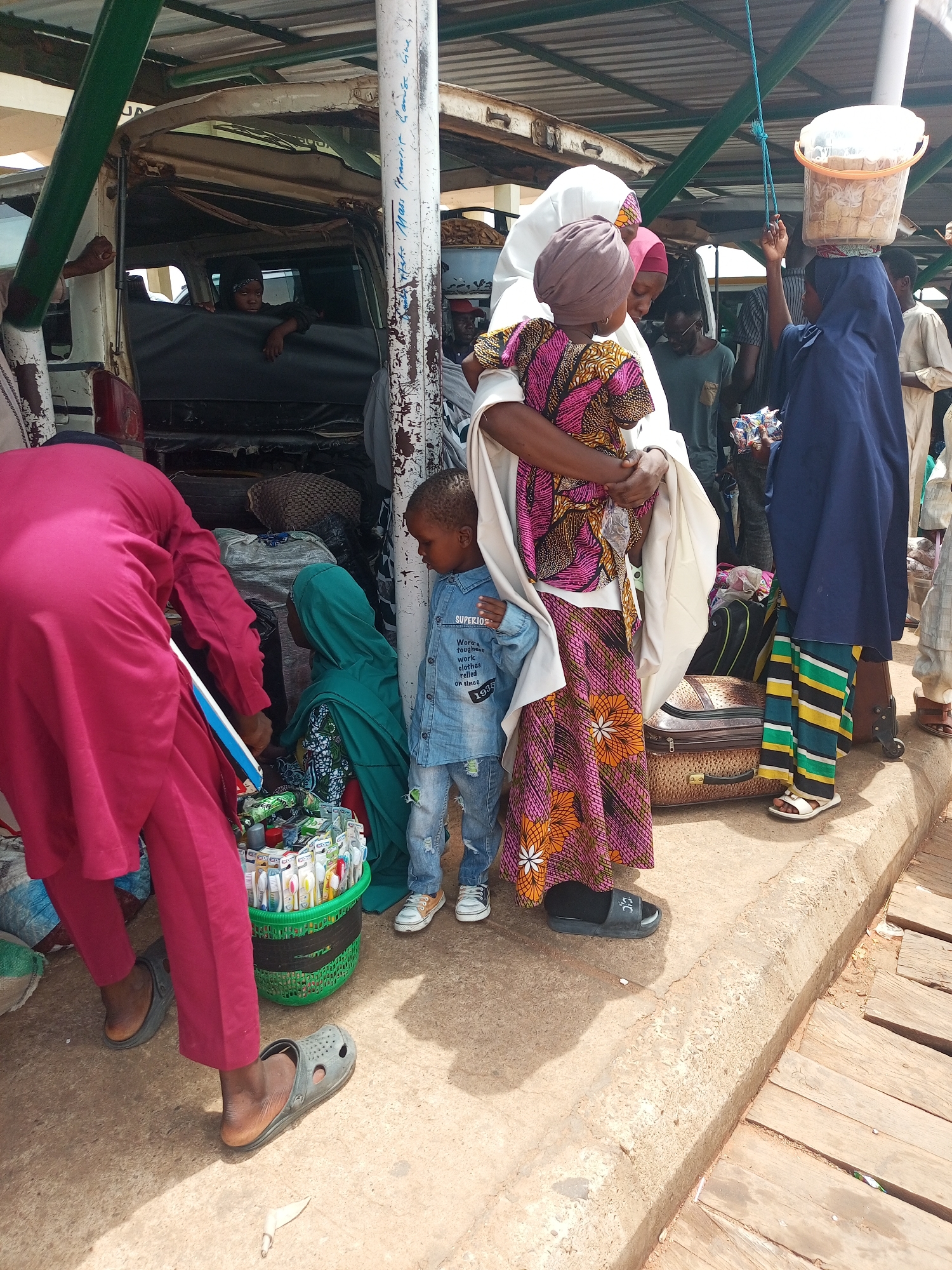 The image depicts a group of people gathered in an urban environment, likely a transportation hub. A woman in a colorful patterned dress and a white head covering is holding a child, possibly a baby, wrapped in a white cloth. Another child stands next to her, wearing a denim jacket. To the left, a person in a magenta robe is bent over, interacting with another individual in a green head covering near a basket filled with various items, possibly for sale. A young girl in a blue hijab is carrying a container on her head. Luggage is placed at their feet, suggesting travel. In the background, a vehicle, possibly a bus or van, is visible with passengers inside. The location appears to be a busy area with a mix of people, vehicles, and activity, suggesting a transportation hub in an unidentified city.
