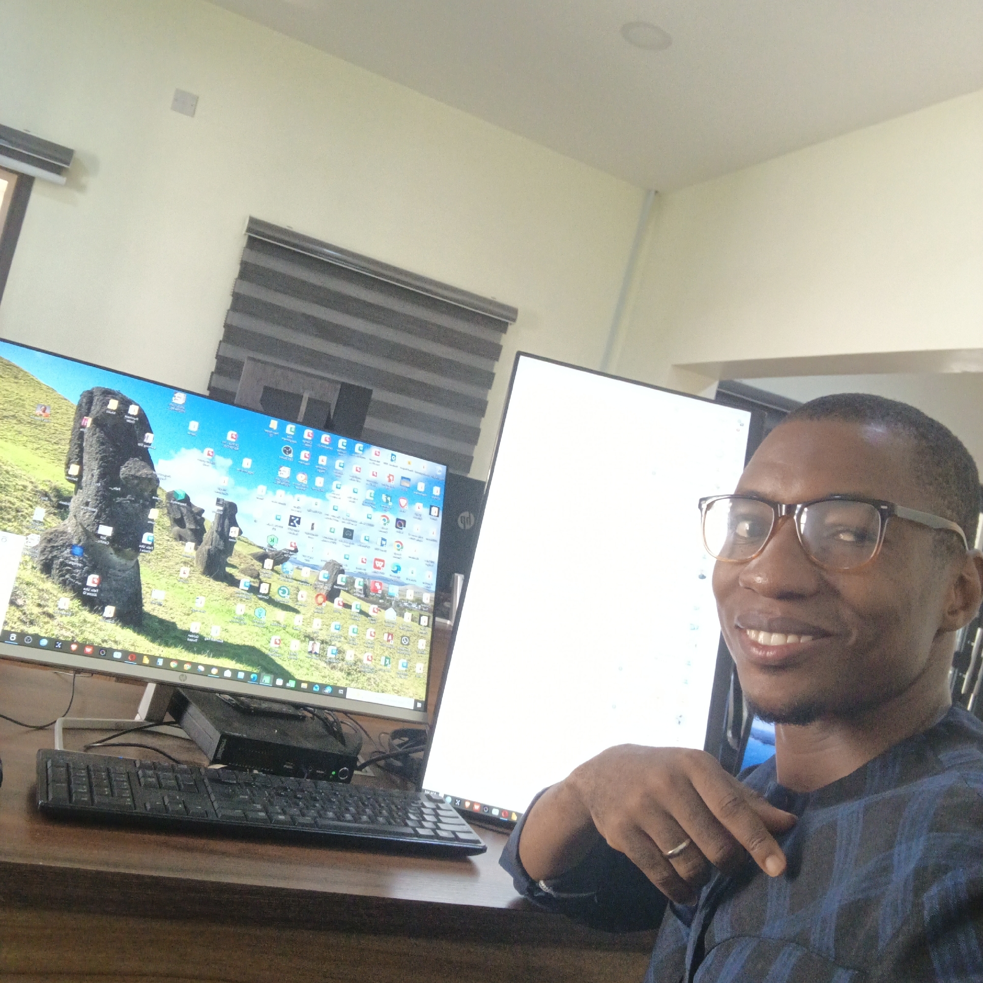 A man wearing glasses and a blue and black patterned shirt smiles and looks at the camera while sitting at a desk with two computer monitors. The monitor on the left displays a desktop background of the Moai statues on Easter Island, along with many program icons. The monitor on the right is mostly white, with a few small, indistinct blue dots visible. A black keyboard is in front of the monitors, and a small black computer tower is placed on the desk behind the keyboard. The man's left hand rests on his chest, and he wears a silver ring on his left ring finger. The scene appears to be an indoor office environment.