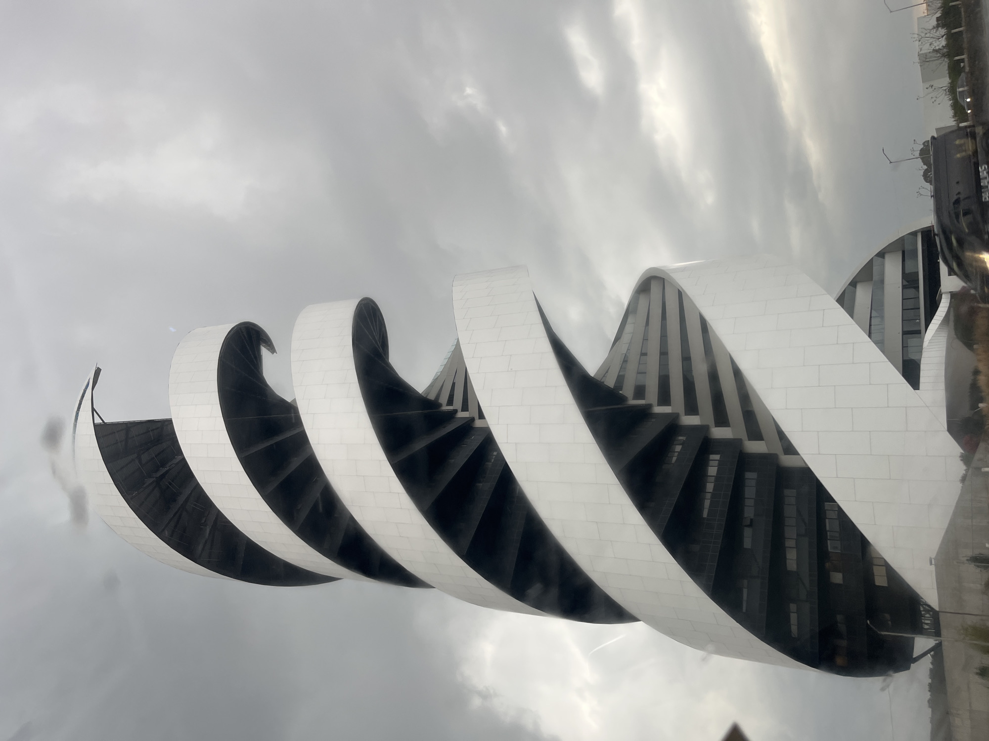 The image depicts the Museo Internacional Barroco in San Bernardino Tlaxcalancingo, Mexico. The museum's distinctive design features a series of white, curved, wave-like structures that appear to spiral or undulate against a backdrop of cloudy, overcast skies. In the spaces between the white curves, dark or shadowed areas suggest internal architecture and possibly staircases or other architectural elements. The building's facade is clad in white tiles. Other buildings and vehicular traffic are visible in the background.