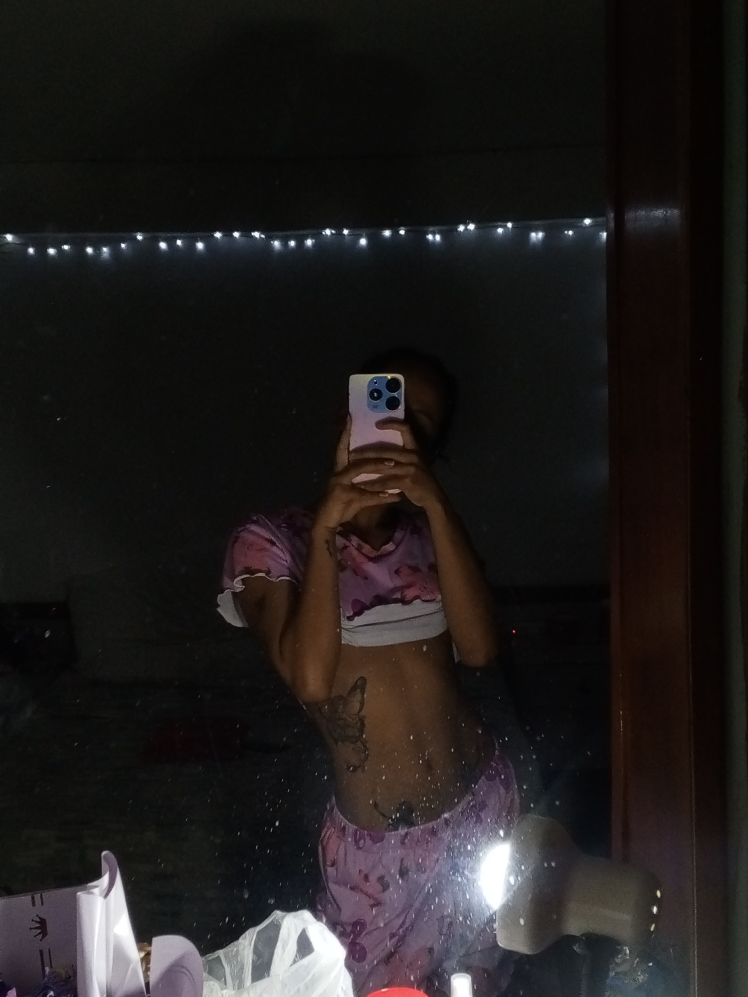 A person is seen from the chest down, taking a mirror selfie in a dimly lit indoor setting, likely in Porlamar, Venezuela. Their face is obscured by a light pink/purple smartphone held up for the photo. They are wearing a patterned pink and purple crop top with white trim, and matching pajama-style shorts or pants. A prominent butterfly tattoo is visible on the left side of their torso, with another smaller tattoo on their abdomen.
The background, reflected in the speckled mirror, is dark. A string of white LED lights stretches horizontally across the upper portion of the scene. To the right, a dark brown wooden door frame is visible. The foreground of the reflection shows various objects: a light-colored box with purple details on the left, and a white plastic bag next to a brightly illuminated object (possibly a flashlight) on the right. The overall impression is of a casual, nighttime moment. There is no visible text in the image.