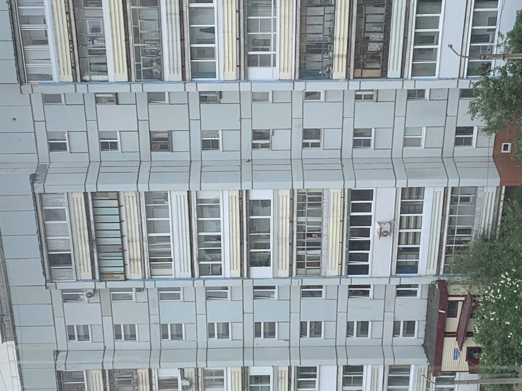 The image depicts the exterior of a large, multi-story apartment building, likely in Chaikovsky, Russia. The building has a symmetrical facade with a repetitive pattern of windows and light-colored panels. The window design alternates between single, small windows and large window sections. The building appears to be constructed of pre-fabricated concrete panels. The overall color palette of the building is dominated by pale grey. The building's facade shows signs of aging and wear. The area in the lower corner of the image shows the top of a smaller building with a red roof. Trees with green foliage are also visible on the side of the image. There is an air conditioner visible on the building's exterior. The image shows no people.