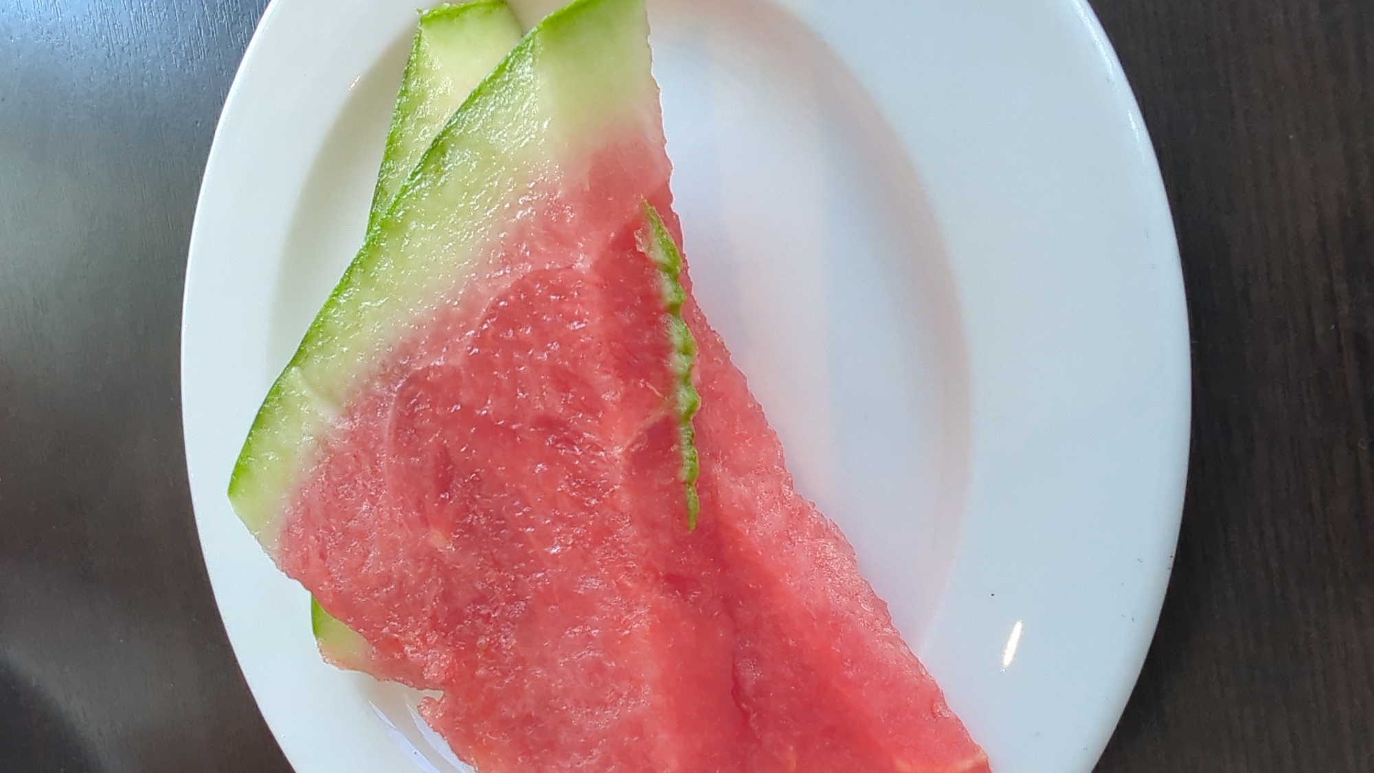 Two slices of watermelon sit on a white plate. The watermelon slices are bright pink with a green rind. The plate is on a dark wooden surface. The scene appears to be an indoor setting. The image does not provide any information about the city of Larkollen, Norway.