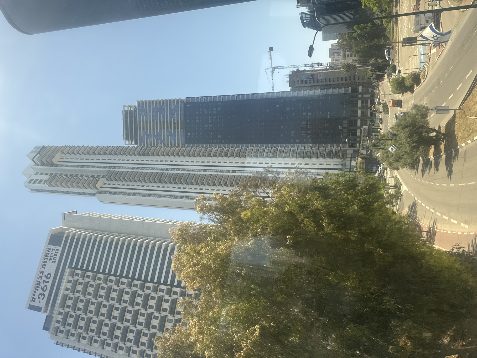 The image depicts a view in Ramat Gan, Israel, featuring a skyline dominated by modern high-rise buildings. The architecture consists of skyscrapers with distinctive designs, including a building marked with the number "3616." A road curves in the foreground, bordered by trees and displaying traffic markings. Israeli flags are visible alongside the road. Construction cranes are present, indicating ongoing development in the area. The weather appears sunny.