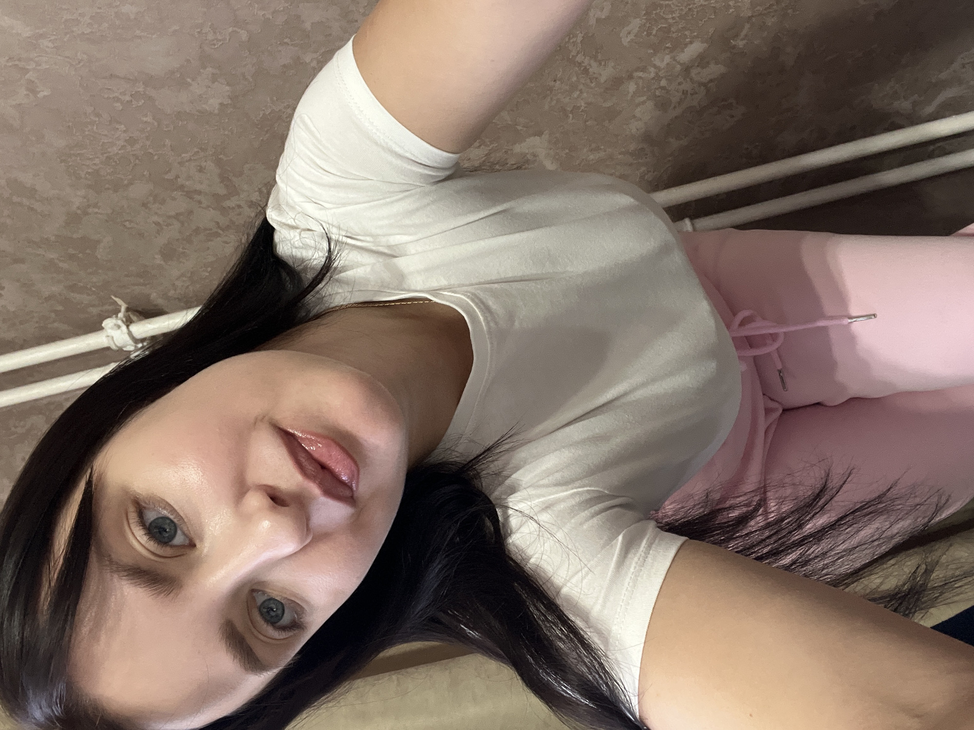 A woman with long, dark hair is positioned at an angle in front of a beige-colored wall. She is wearing a white t-shirt and pink sweatpants. A white radiator is visible in the background.
Location: The image does not offer enough clues to definitively conclude that the image was taken in Марковский, Russia.