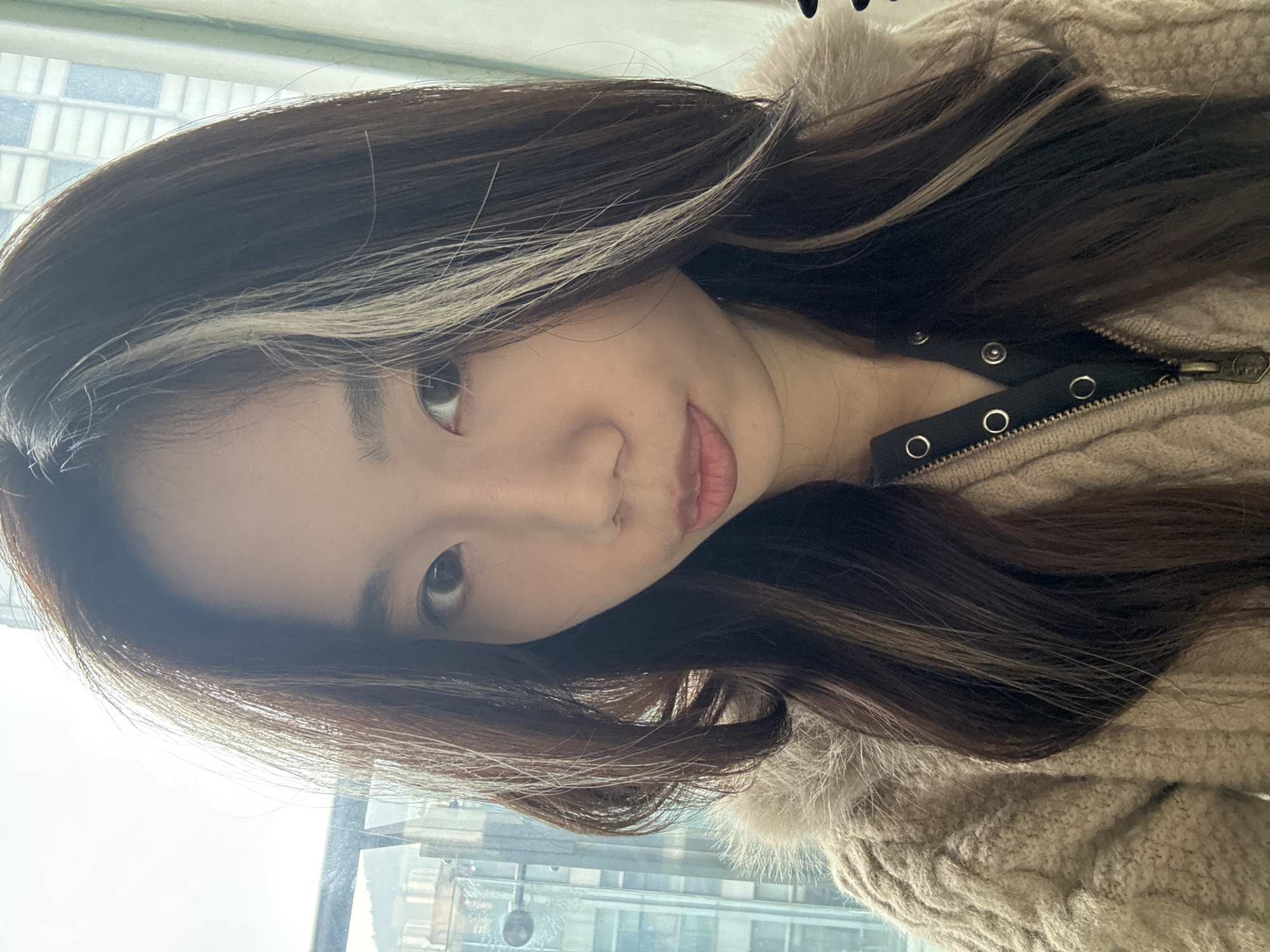 The image depicts a female of apparent East Asian ethnicity, captured from the upper chest to the top of her head, centered in the frame. She is located indoors, likely within a high-rise building in the Nan'an District, China, with an urban cityscape visible through a large window behind her.
The individual has dark brown hair, styled long and parted, featuring distinct lighter brown or blonde streaks on both sides of her face, primarily above the temples and extending downwards. Her eyes are dark, and her mouth is closed with lips slightly rounded forward.
She is wearing a light brown, possibly beige, cable-knit or textured sweater with a visible faux fur collar around the neckline and shoulders. Beneath this, a dark garment, potentially a shirt or an inner layer, is visible at the V-neck opening. This garment features several silver-colored snap buttons and a metal zipper, which is partially unzipped.
The background shows a clear daytime sky and the upper sections of multiple modern, glass-facade high-rise buildings, indicative of an urban environment. A window frame or ledge is partially visible on the left side of the background. The individual is looking directly towards the camera.