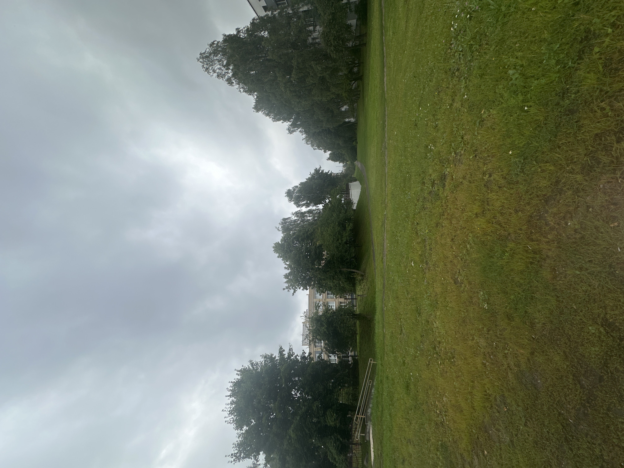 A wide-angle, eye-level shot captures a residential courtyard in Minsk, Belarus under an overcast sky. The foreground is dominated by a lush green lawn, with a patch of drier, yellowish grass on the left. A dirt path runs along the bottom left edge of the frame. In the middle ground, a paved pathway winds through the grass, flanked by trees. To the left, a small ramp with yellow handrails leads up to a fence. Behind the trees and the path, a multi-story apartment building with light-colored facades and numerous windows is visible. Further to the right, more trees line the landscape, with another apartment building on the far right edge of the image. The overall scene depicts a typical urban green space in a residential area.